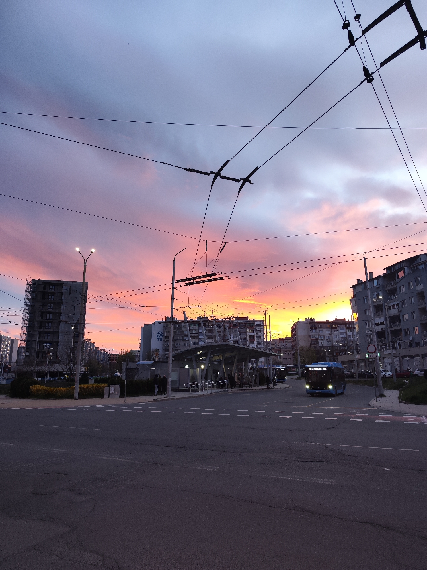 The image shows an urban scene in Burgas, Bulgaria, at twilight. The sky is a mix of colors, ranging from orange and yellow near the horizon to pink and purple higher up. Silhouetted buildings line the horizon, including a gray building under construction on the left with scaffolding visible. Overhead trolley wires stretch across the image.
In the foreground is a road intersection with zebra crossings. A blue bus is approaching the intersection. A covered bus stop is located on the right side of the road, occupied by a few people. Bus stop has an inscription that says: "10 Пг Роса - СГ". Yellow flowers are planted in a green area near the bus stop. Cars are parked in the background along the street. The image captures an everyday scene in the city during the transitional period of sunset, with a focus on public transportation infrastructure.