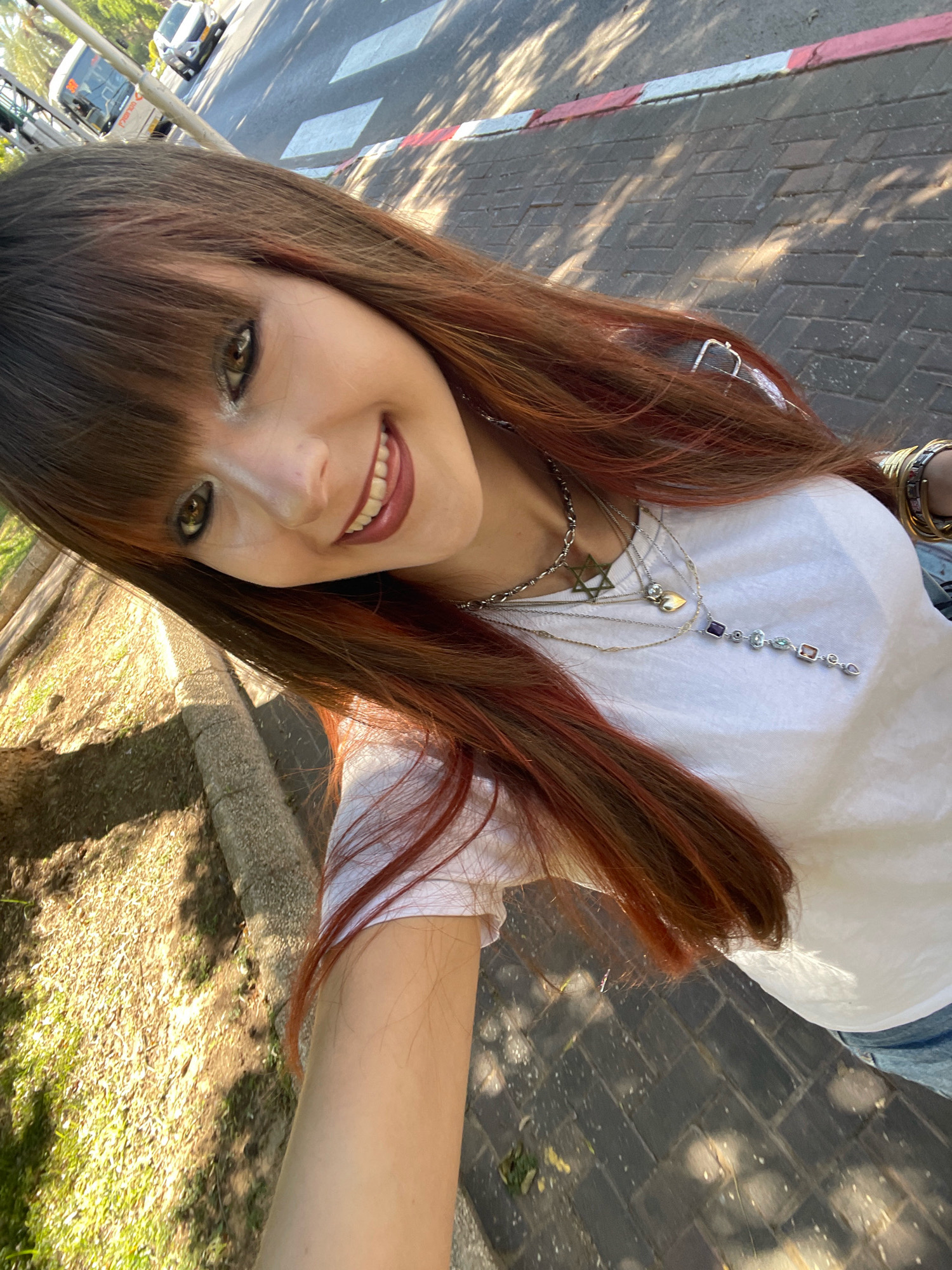 The image shows a young woman taking a selfie outdoors in Raanana, Israel. She has long, auburn hair with bangs and is wearing a white t-shirt and blue jeans. She is smiling and has makeup on, including dark eyeliner and lipstick. She is wearing several necklaces, including one with a Star of David pendant and another with colorful gemstones. She is also wearing bracelets on her wrist.
The background shows a sidewalk with patterned paving stones and a tree-lined street. There is a crosswalk visible in the distance, and a bus and a car are on the road. The sun is shining, casting shadows on the ground. The woman is standing near a grassy area with a stone curb. The overall impression is of a casual outdoor scene in an urban environment.