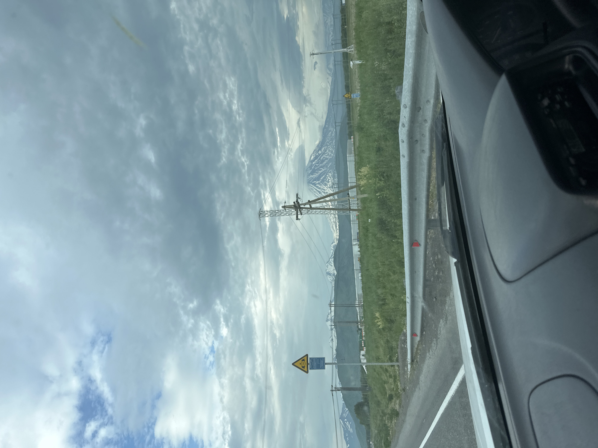 The image shows a view from inside a vehicle, looking out at a cloudy sky and a mountainous landscape. In the foreground, the dashboard of the car is visible. A road with a white line runs along the left side of the frame, with a metal guardrail to the right. There are power lines and utility poles scattered throughout the middle ground. A triangular yellow warning sign with an image of a person and a smaller blue sign below it is mounted on a pole to the left of the road. Further in the distance, snow-capped mountains are visible under a heavy cloud cover. There are no people or animals visible in the image. The scene appears to be in a rural or semi-rural area, likely on the outskirts of a settlement. The overall impression is of a somewhat overcast day in a natural setting with infrastructure elements. The presence of the warning sign and power lines suggest human activity in the vicinity. Given the context of Кеткино, Russia, this scene likely depicts a view of the surrounding natural terrain and infrastructure in that region.