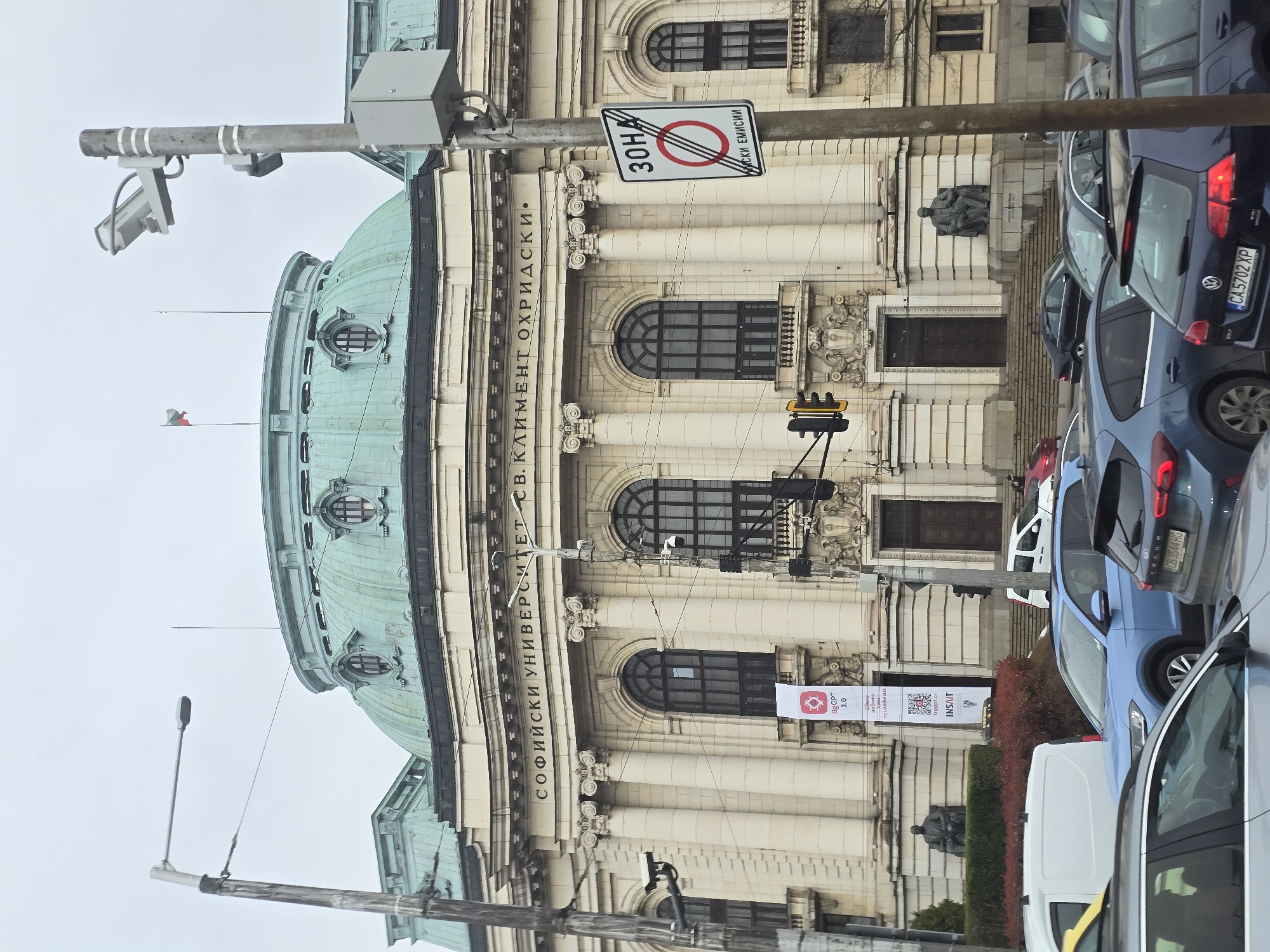 The image is of Sofia University St. Kliment Ohridski in Sofia, Bulgaria. The building is beige with a green dome. There is a sign that reads "СОФИЙСКИ УНИВЕРСИТЕТ СВ. КЛИМЕНТ ОХРИДСКИ•". There is also a 30 km/h speed limit sign. A banner reads "BgGPT 3.0 новото bggpt.al INSAIT". There are cars parked in front of the building, and a statue on the building.