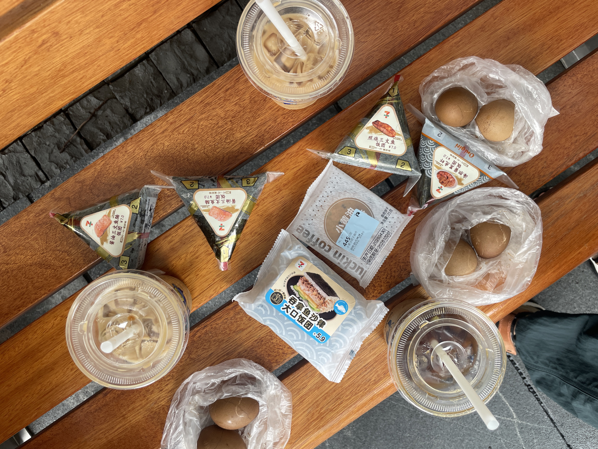 On a wooden bench, arranged are three cups of iced coffee with straws, four triangular onigiri rice balls in branded wrappers, two bags each containing two brown eggs, a Luckin Coffee packaged cookie, and a 7-Eleven packaged rice ball. The onigiri varieties include salmon and prawn. The background includes gray brick, suggesting an urban environment. The location is likely in Tianhe District, China, given the presence of Luckin Coffee and 7-Eleven branding. A person's leg in dark pants is visible on the lower right.
