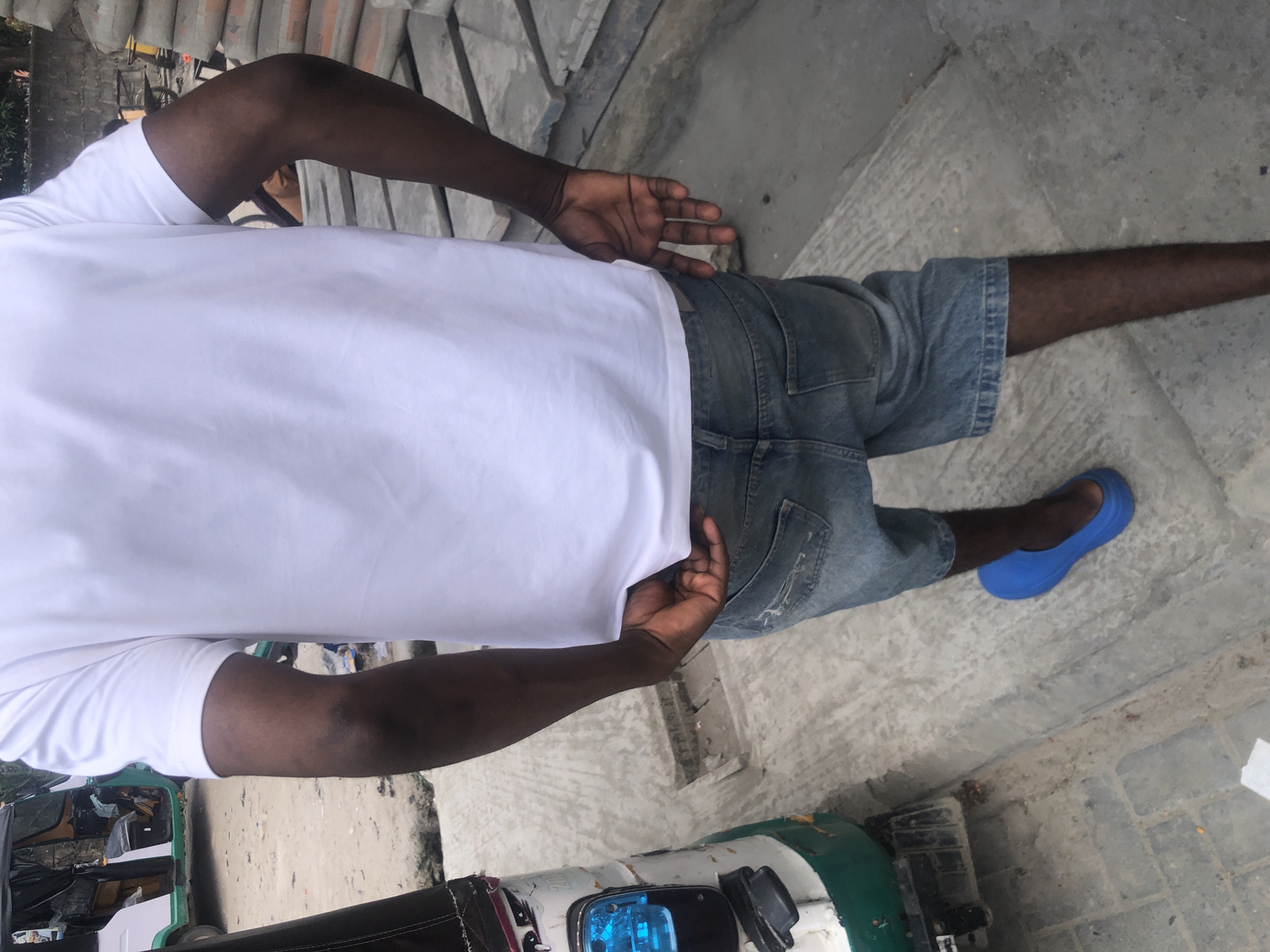 A medium shot shows the back of a Black male wearing a white t-shirt and denim shorts. His left hand is tucked into his shorts pocket. He is wearing a single blue rubber clog on his left foot. The background shows a concrete surface, a pile of stacked concrete slabs, and a portion of a green and white three-wheeled vehicle. This scene appears to be located in the city of Ado, Nigeria.