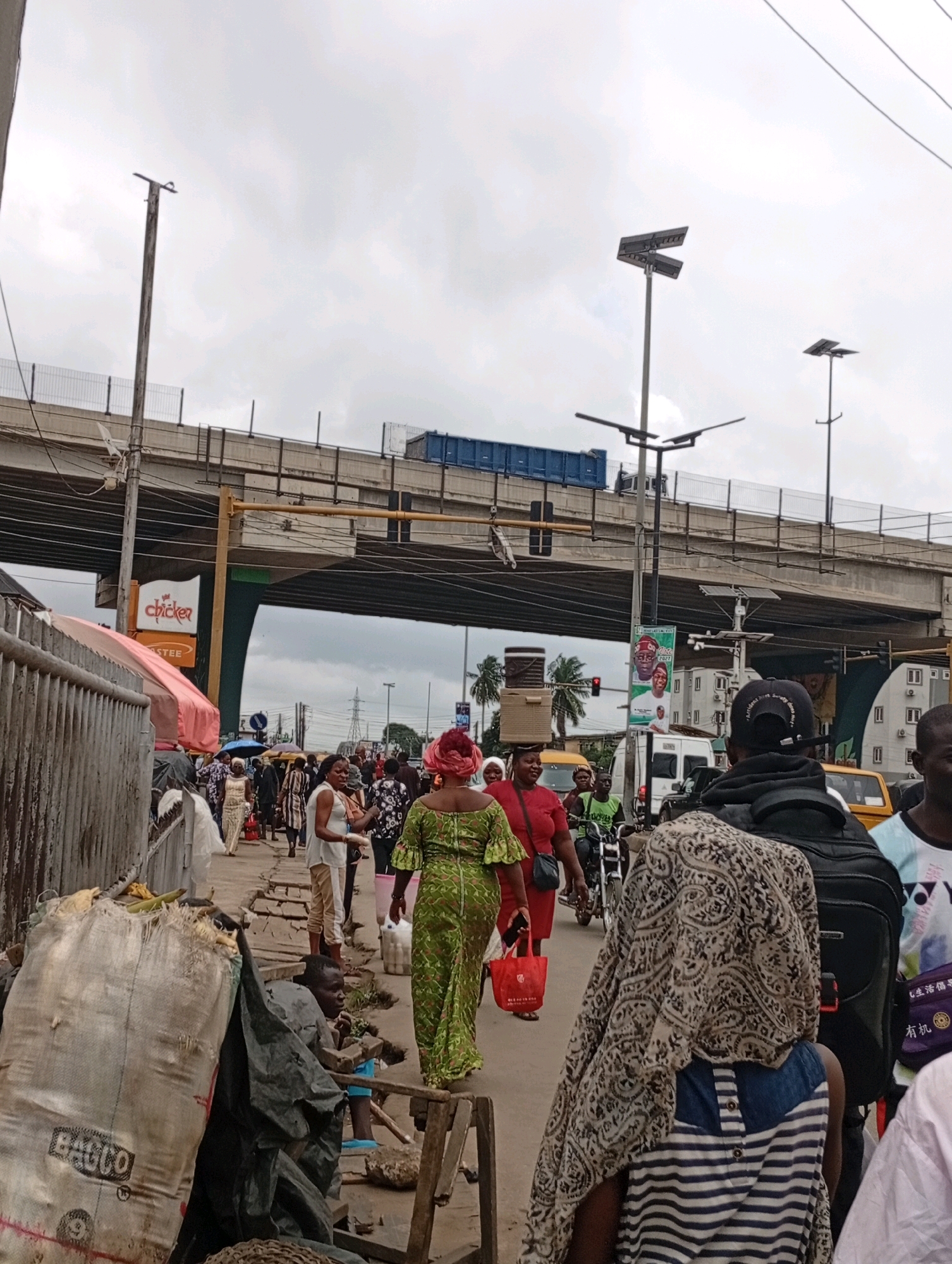 The image depicts a busy street scene under an overcast sky, likely in Lagos, Nigeria, with an elevated highway system dominating the background. In the foreground, a group of people navigate a crowded walkway alongside a corrugated metal fence. One person, partially obscured, is seated next to a large, off-white woven bag labeled "BACOO". To their right, a person in a green patterned dress walks away from the camera, carrying a red shopping bag. Other individuals in the mid-ground are dressed in various casual and traditional clothing, some carrying items or interacting. Behind them, vehicles, including a yellow taxi and a motorcycle, are visible. The elevated highway has a blue shipping container visible on it. Several streetlights, some with solar panels, line the area. The overall environment suggests a bustling urban market or commercial district.