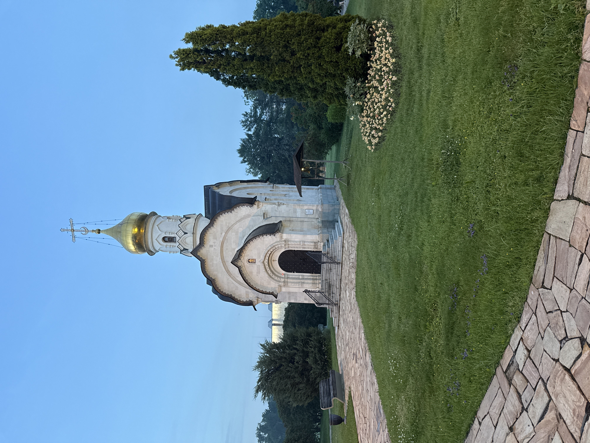 The image shows a small, white, Orthodox chapel with a golden dome and a cross on top, situated in a park-like setting. The chapel has a dark, arched doorway with steps leading up to it. To the left of the chapel, there is a stone pathway, a wooden bench, and a trash receptacle. To the right of the chapel, there is a tall evergreen tree, a bed of light-colored flowers, and a wooden structure that appears to be a small shelter. The background consists of lush green trees and a clear, pale blue sky, suggesting it is either dawn or dusk. A hint of buildings can be seen in the distance behind the trees on the left.
This chapel is likely the Church of the Deposition of the Robe in the Kremlin. The Church of the Deposition of the Robe is a cathedral in the Moscow Kremlin, Russia.