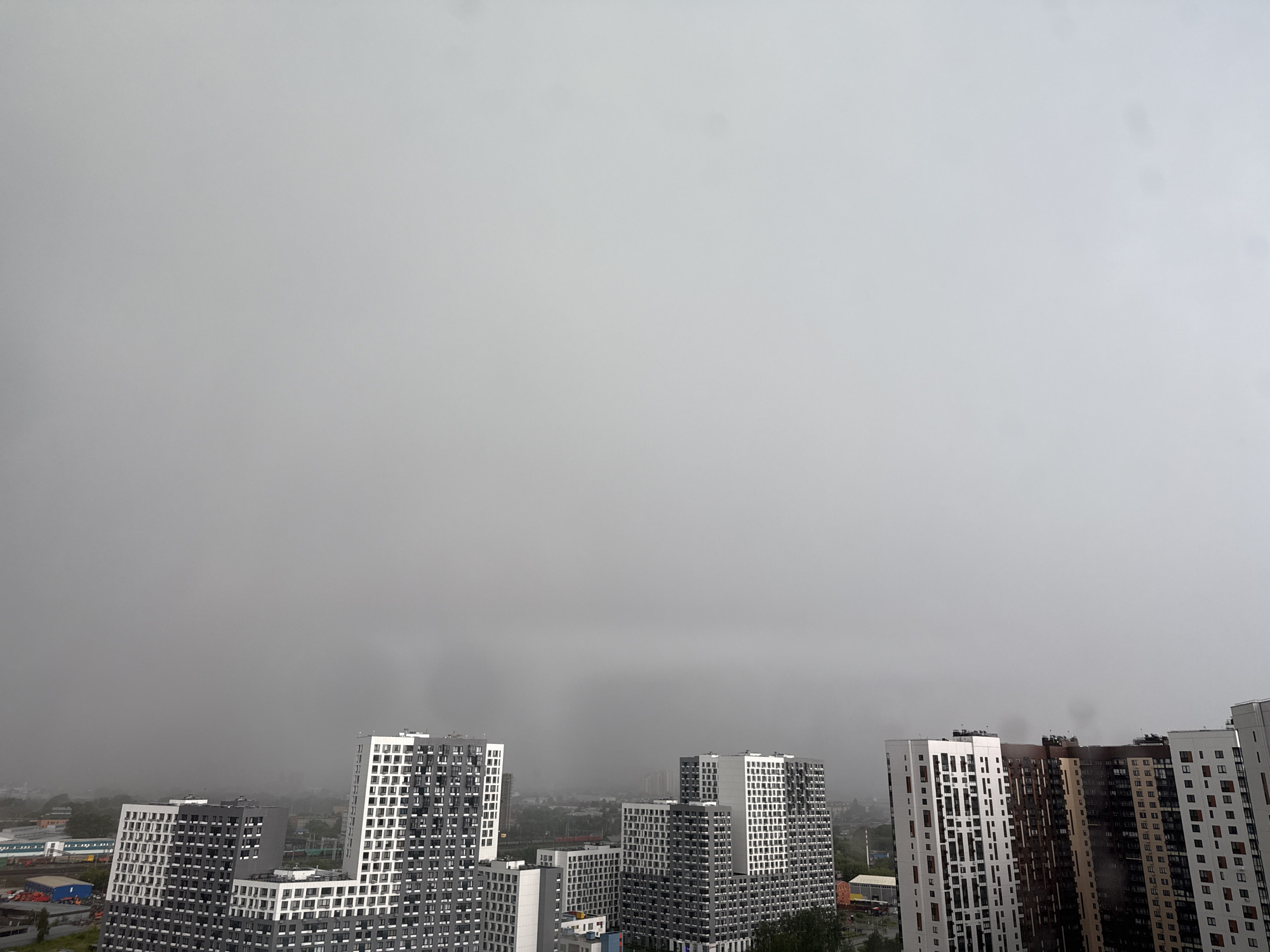This is a wide, aerial shot of a modern urban landscape in Lyubertsy, Russia. The sky is overcast and a thick, gray haze obscures the distant horizon, giving the impression of either fog or heavy pollution. In the foreground and midground, several tall, contemporary apartment buildings are visible. These buildings are characterized by their white and dark gray facades, with repetitive patterns of windows. Some of these buildings are partially obscured by the atmospheric haze. To the left, industrial buildings and railway tracks are discernible, suggesting a mixed-use or industrial area adjacent to the residential developments. Vegetation, such as trees, is also present, particularly towards the bottom of the frame, suggesting some green spaces within the city. The overall scene depicts a dense urban environment under a muted, gray sky.