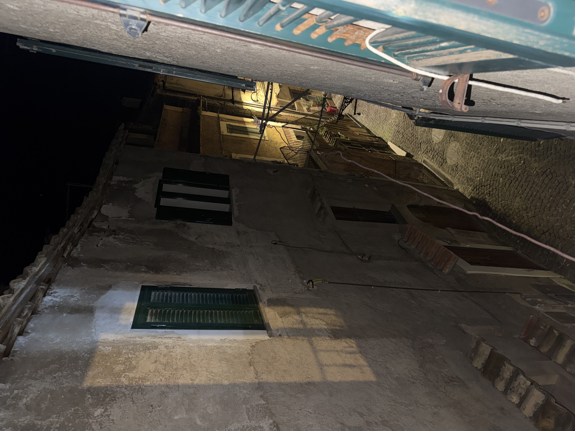 The image shows a narrow street view looking up in Castelnuovo di Porto, Italy, at night. Tall, old buildings flank the street, featuring weathered stucco walls, dark green shutters on windows, and tile rooftops. Electrical wires run along the walls. Overhead, a green metallic awning is visible. The scene is dimly lit, with some light emanating from the upper windows of the buildings, illuminating the cobblestone pavement.