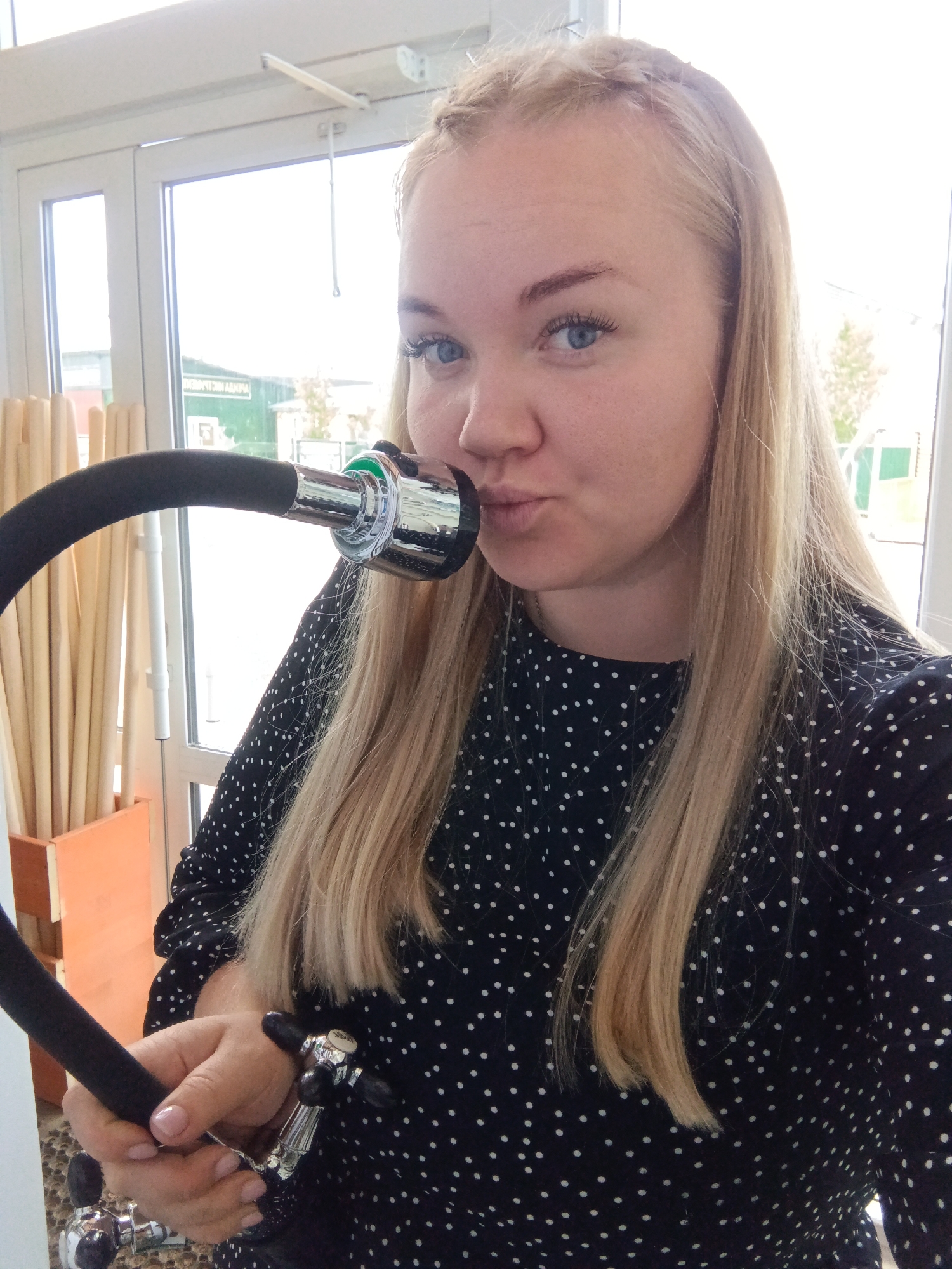 A young woman with long blonde hair, fair skin, and blue eyes is captured in a playful pose indoors. She wears a dark, possibly navy blue or black, long-sleeved top or dress featuring small white polka dots. Her hair is styled with a subtle braid along the top of her head.
She is holding a flexible black hose, which terminates in a chrome metal nozzle with a green ring accent. The woman has brought this nozzle close to her face, puckering her lips as if to kiss or blow into it, while looking directly at the camera with an engaging, lighthearted expression. Her right hand is visible in the lower right of the frame, holding another chrome fitting, suggesting she is interacting with a plumbing fixture or a similar display item.
The background is dominated by a large, bright window or glass door, indicating a daytime setting. Through the glass, part of an outdoor scene is visible, including a building with a green facade. Reflections on the window show reversed Cyrillic text, which appears to spell "ДВЕРНОЙ АЛЬЯНС" (DVERNoy AL'YANS), likely a store or business name. To the left, an indoor display holds numerous upright wooden sticks or dowels in a wooden and orange container. The overall setting suggests a retail environment, such as a hardware store or showroom, consistent with the provided location context of Фоки, Russia.