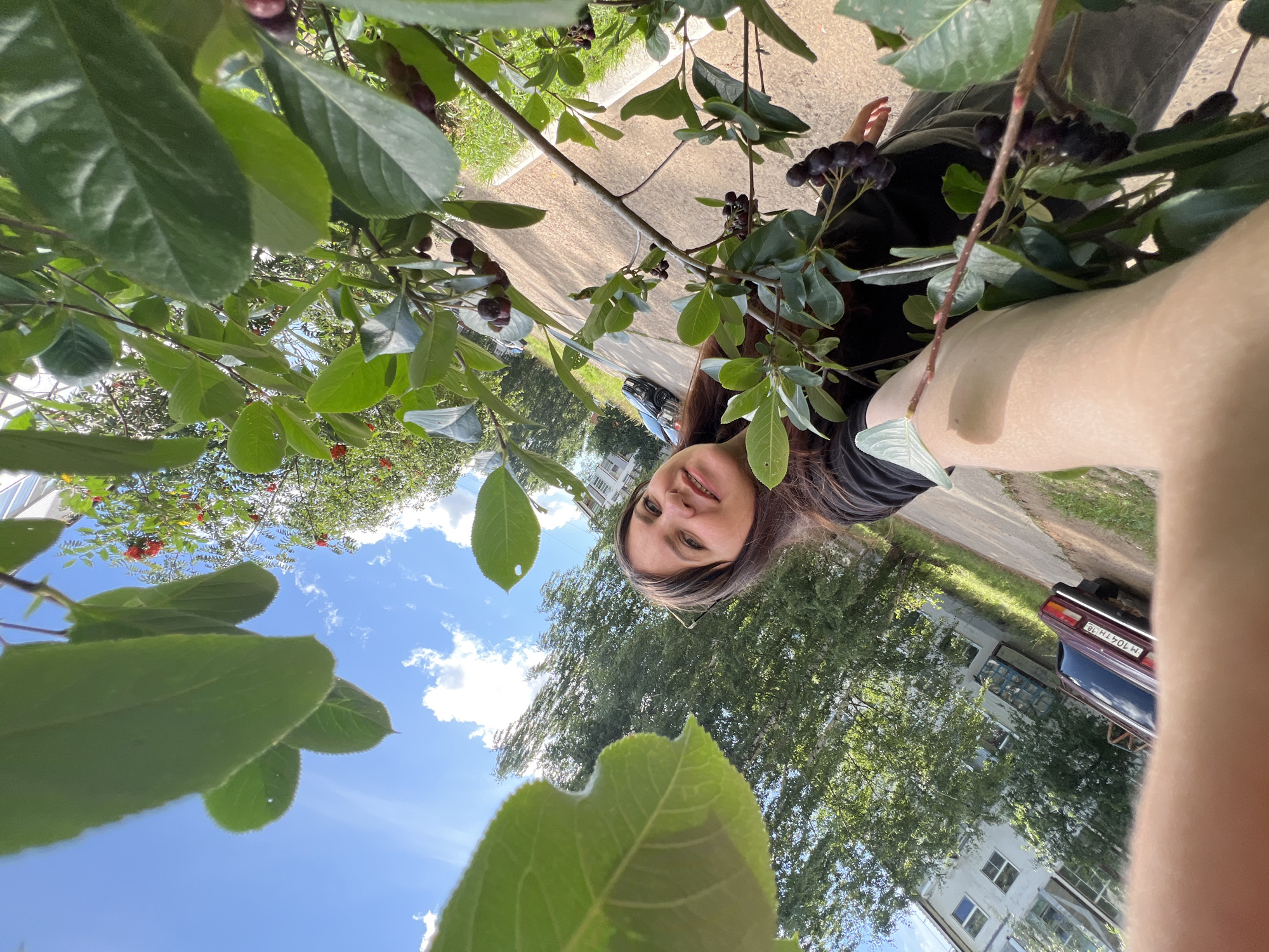 A woman is taking a selfie in Noviy, Russia, near a residential building. She is smiling at the camera, with a branch of aronia berries in the foreground partially obscuring her face and arm. The woman is wearing a black t-shirt and grey pants. In the background, a parked red car is visible next to a paved path and a grassy area. Trees and a multi-story apartment building are also present in the scene. The sky is blue with scattered clouds.