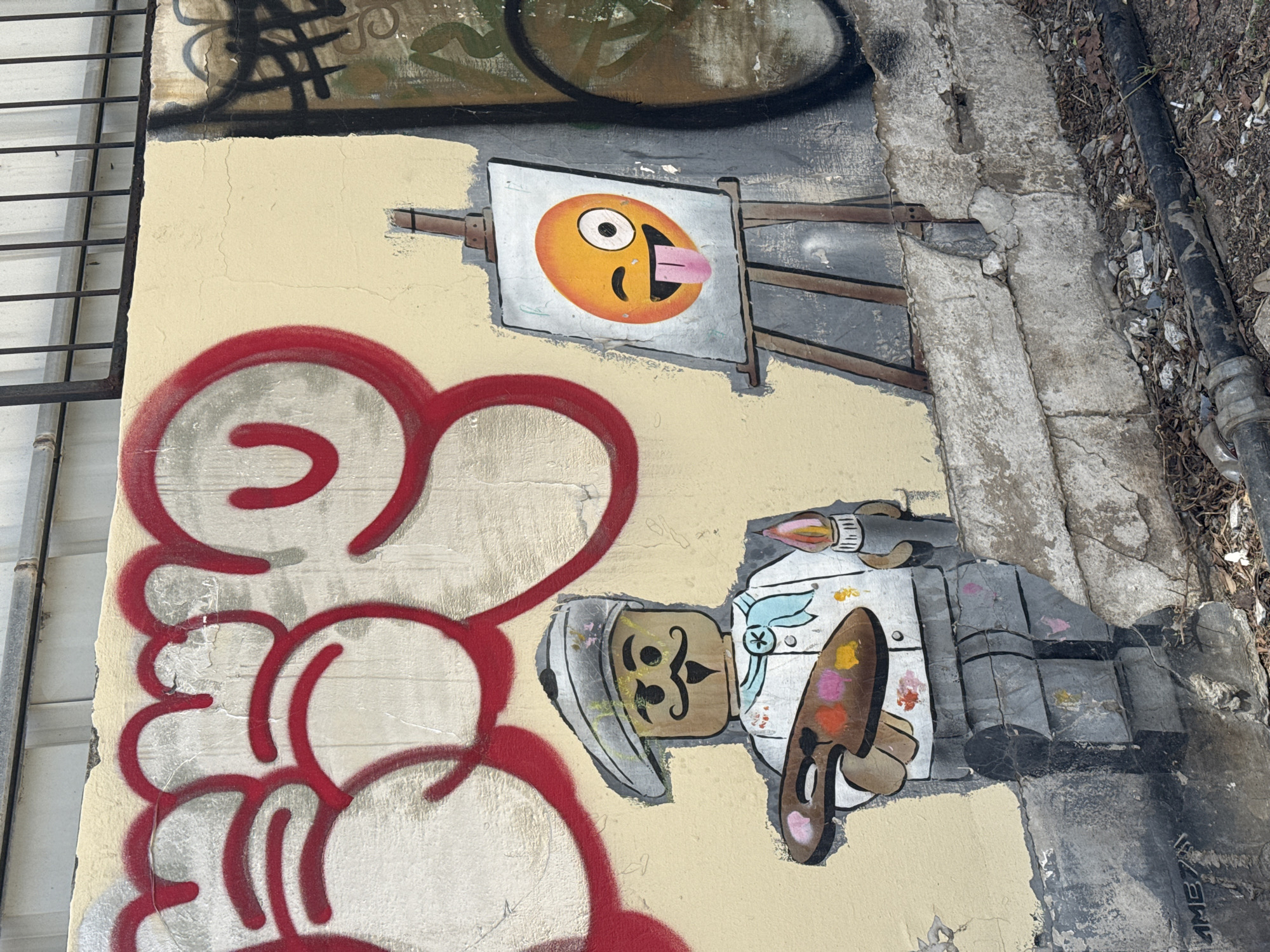 The image displays a segment of a weathered, light-colored wall adorned with multiple layers of street art in an urban environment. The central artwork features a cartoonish character, reminiscent of a chef or baker, dressed in a white uniform, chef's hat, and light blue bandana. This character, sporting a prominent mustache, holds a brown artist's palette with dabs of colorful paint (pink, yellow, orange) and a paintbrush.
Above and slightly to the right of the character, an easel presents a painting of a playful, yellow-orange emoji face. This emoji has one large, wide-open eye and a smaller, crescent-shaped eye, with a pink tongue comically sticking out.
To the left of the main artwork, bold red graffiti in rounded, bubble-like shapes or letters extends downwards. Above the primary graffiti, an older, darker tag is partially visible but largely illegible. The wall itself shows signs of wear, including cracks and some peeling paint.
The surroundings include a vertical metal grate or fence on the far left. To the right, the scene transitions to a cracked concrete curb, followed by a strip of dirt, small stones, and a black pipe. The natural daylight suggests the photograph was taken during the day. A small, black signature or tag, possibly "JMB", is faintly visible near the bottom right of the main graffiti piece.