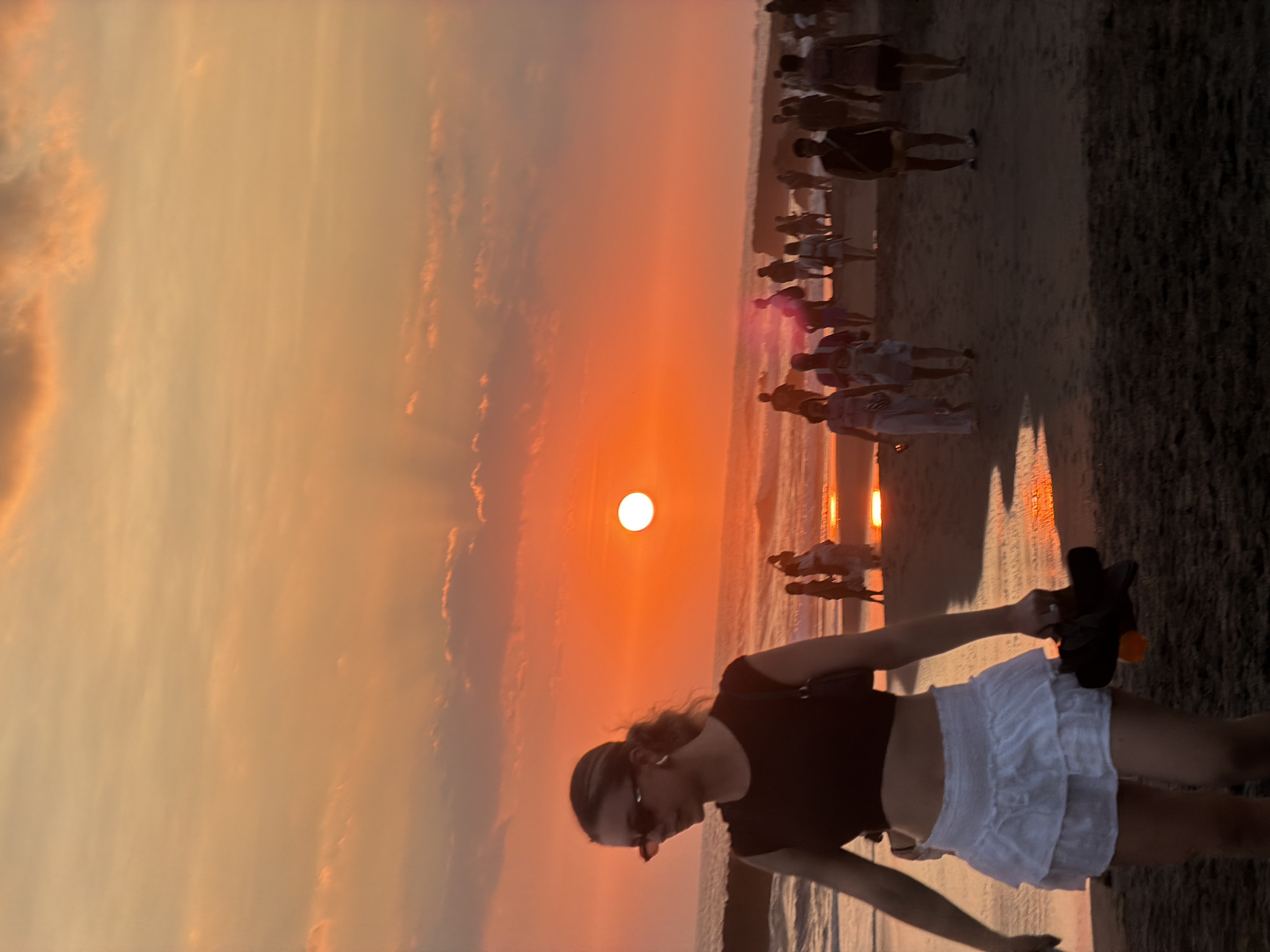 A young woman with blonde hair pulled into a ponytail, wearing sunglasses, a dark crop top, and a white ruffled skirt, walks on a sandy beach in front of a sunset. She is holding a pair of dark sandals with orange soles in her right hand. In the background, other people are walking along the beach and in the ocean. The sun is setting over the ocean, casting a warm orange glow across the sky and reflecting on the wet sand. The location appears to be Tanah Lot, in the city of North Kuta, Indonesia, a popular tourist destination known for its stunning sunsets.