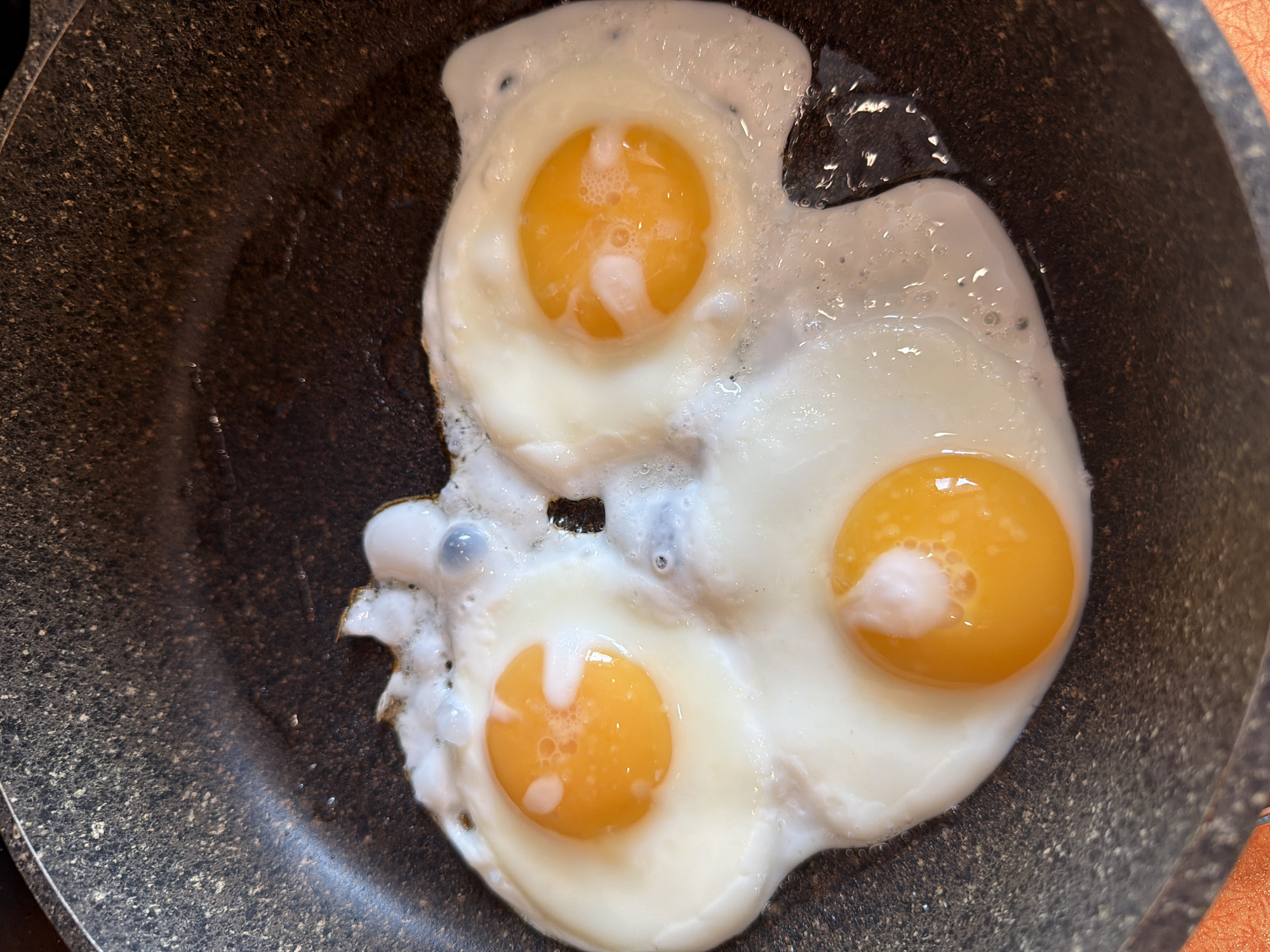 Three fried eggs are cooking in a dark, speckled frying pan. The eggs are sunny-side up, with bright orange yolks and glistening whites. The pan appears to be on a stovetop, as indicated by the heat emanating from the cooking eggs. There are no people or other objects visible in the image. The scene is tightly cropped, focusing solely on the food and the pan. The image does not provide any information related to the city of Solikamsk, Russia, or any specific location.