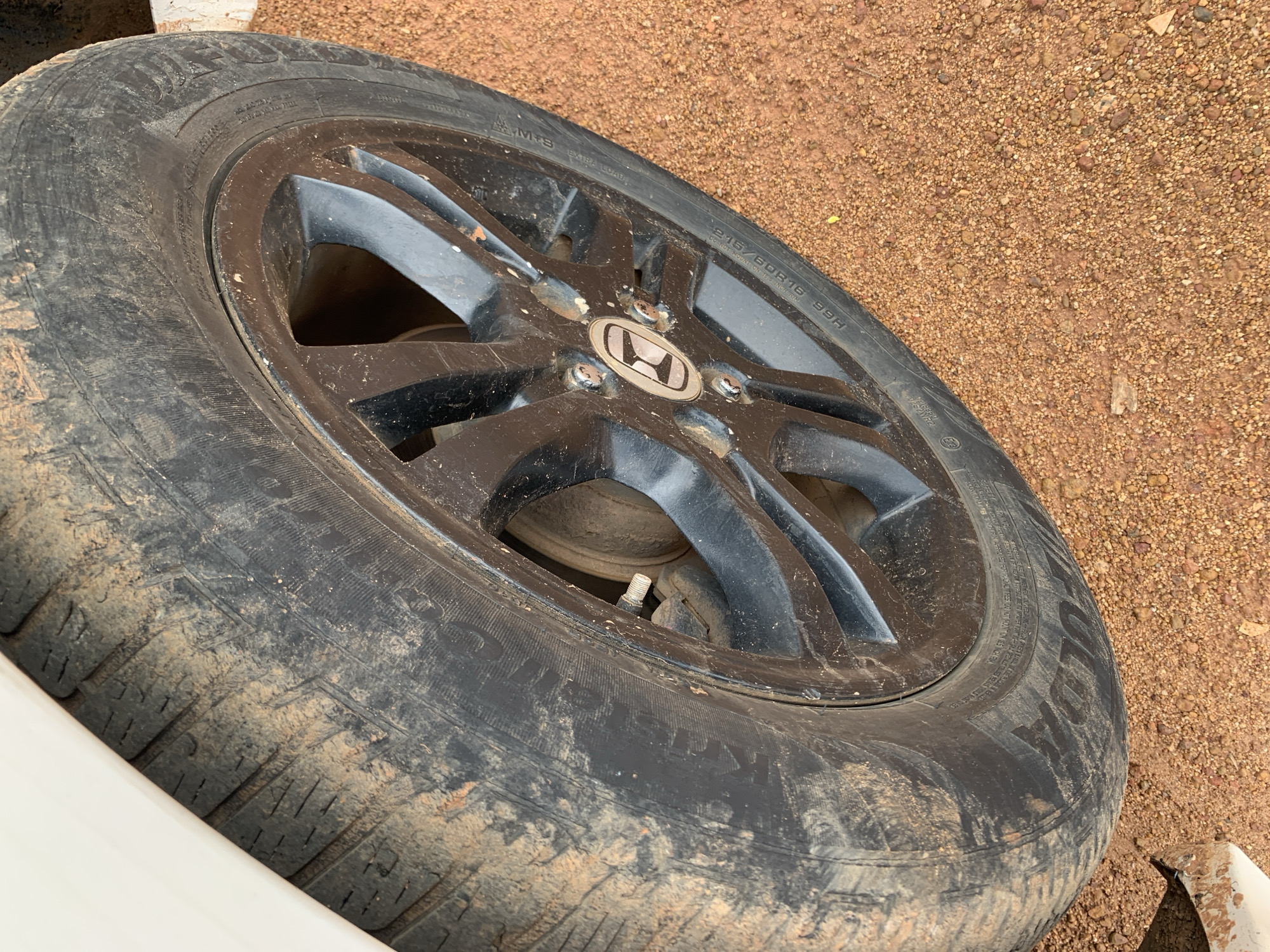 The image shows a close-up of a muddy tire and a black alloy wheel of a vehicle, likely a car or SUV, parked on a reddish-brown dirt surface. The tire is partially obscured by dirt, suggesting it has been driven on an unpaved road. The alloy wheel has a multi-spoke design and appears to have some scuff marks. A circular emblem with a stylized "H" is visible in the center of the wheel. The ground is covered in small stones and pebbles. The lighting suggests it is daytime. This scene is consistent with an outdoor environment in a place like Jos, Nigeria, where dirt roads are common. There are no people or discernible actions depicted in the image.