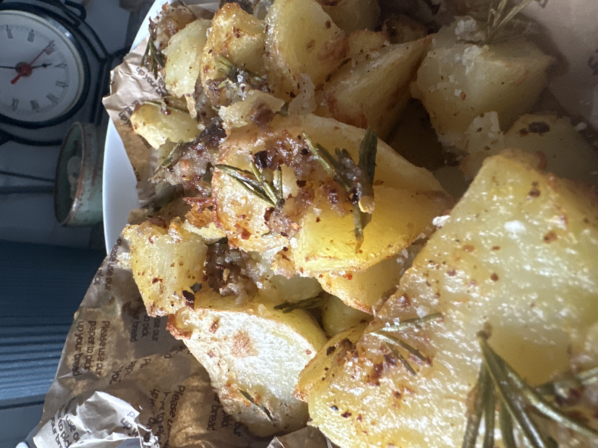 A close-up shot showcases a bowl of golden-brown roasted potato wedges, seasoned with herbs and spices, resting on crumpled parchment paper. The potatoes exhibit a crisp exterior and a fluffy interior, with visible rosemary sprigs and flecks of red chili. In the background, a vintage alarm clock with Roman numerals and a small ceramic bowl are partially visible, hinting at a kitchen or dining area setting. The overall lighting is warm and inviting, highlighting the texture and color of the food. No specific location in Macclesfield, United Kingdom can be identified from the image.