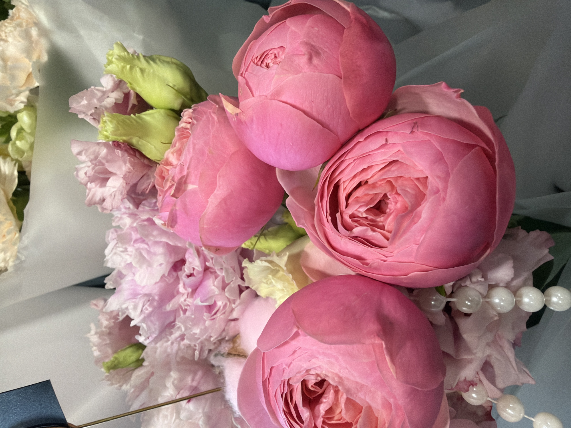 A close-up shot features a bouquet of pink roses and other delicate pink flowers, possibly peonies or carnations. The roses, in various stages of bloom, dominate the foreground. Some are fully opened, revealing their intricate petal structure, while others are tightly budded. A strand of pearls dangles among the flowers, adding a touch of elegance. The background is a soft, neutral gray, possibly depicting a draped fabric or a blurred backdrop. The overall impression is one of softness, romance, and refined beauty. There is no indication of a specific location within a city.
