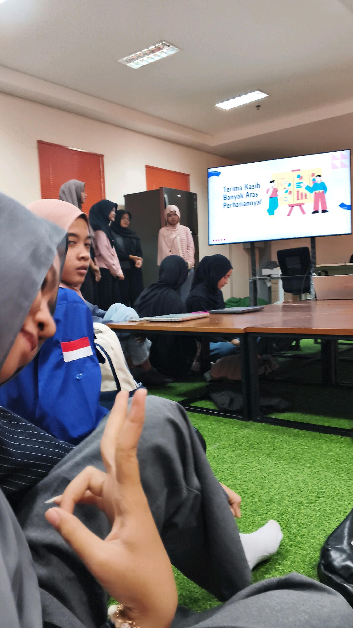 In a room with a green carpeted floor, a group of people are seated and standing, facing a large screen. The screen displays "Terima Kasih Banyak Atas Perhatiannya!" which translates to "Thank you very much for your attention!" in Indonesian. The people are mostly women, many wearing hijabs. In the foreground, a person wearing a grey hijab and a blue jacket with a red and white patch is visible, with their hand making an "OK" gesture. They are seated on gray pants. Behind them, other individuals are seen in various poses, some looking towards the screen, others engaged in conversation. A table with laptops is in the middle ground. The overall atmosphere appears to be a presentation or a meeting. The location is identified as Lohbener, Indonesia.