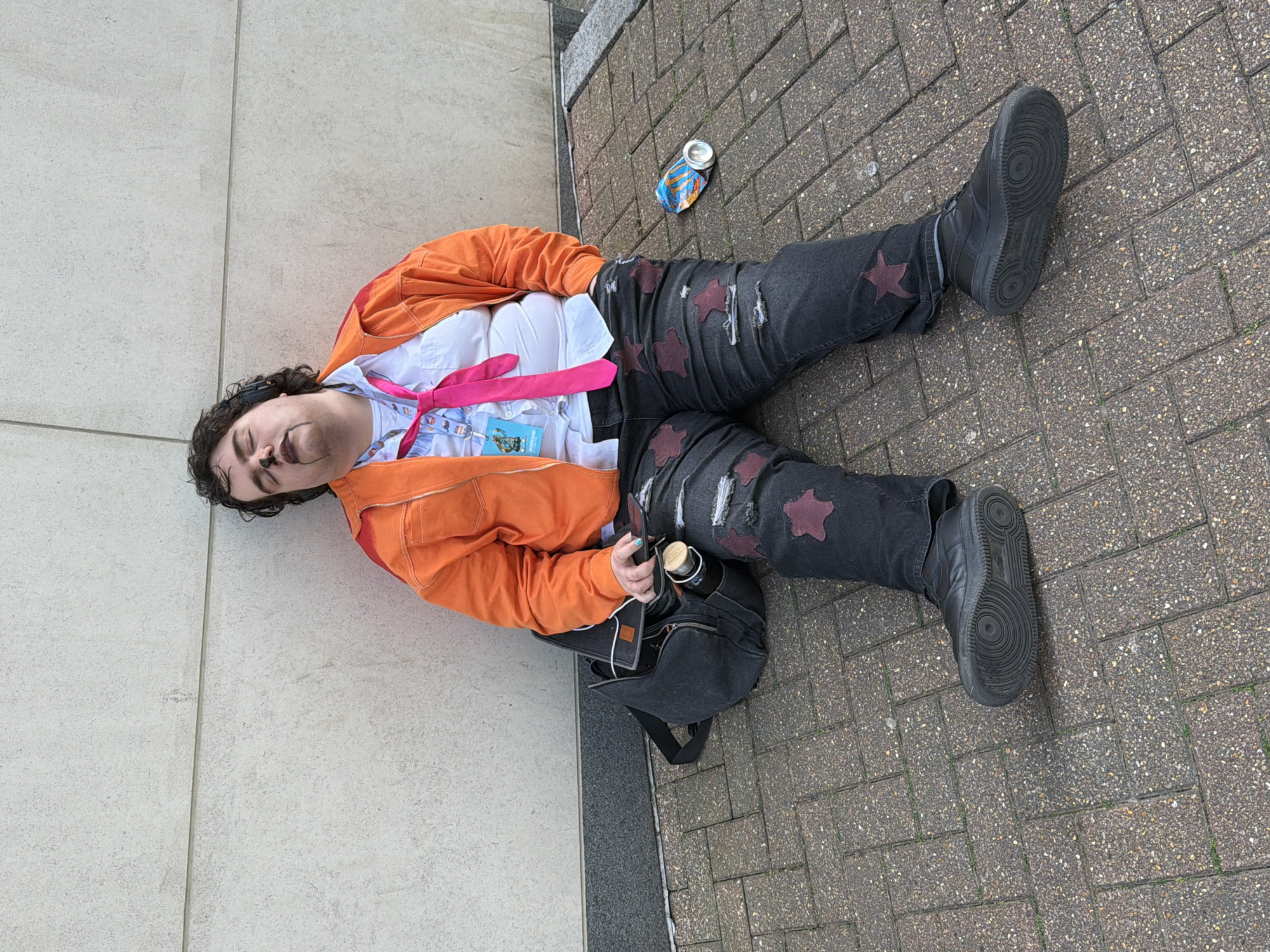 A person lies on the ground on a brick sidewalk next to a building in London, United Kingdom. The person has dark curly hair and is wearing an orange jacket over a white shirt and black jeans with star-shaped patches. A pink tie is draped around their neck, and an identification badge is attached to their shirt. A crumpled can sits near the person's legs. The person is lying on their left side with their head resting against the building. A black backpack with a water bottle inside is positioned near their torso. They wear black sneakers.