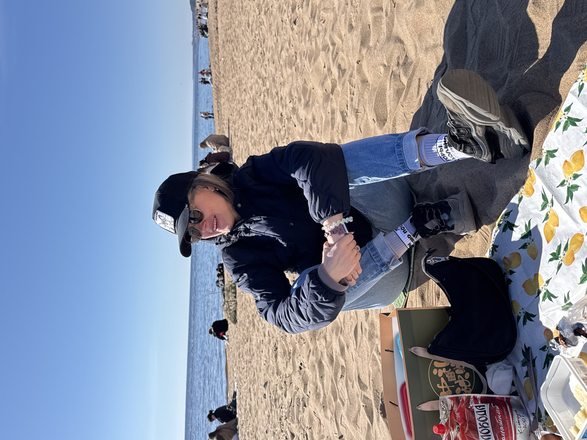 The image depicts a person sitting on a sandy beach in Komarovo, Russia. The sky is clear and blue, and the sea is visible in the background. Other people are scattered along the beach. The individual is wearing jeans, a dark jacket, sneakers, patterned socks, a black cap, and sunglasses. They are seated on a patterned blanket next to a picnic setup including food containers, a paper bag, and a purse. The picture appears to be taken during a sunny day.