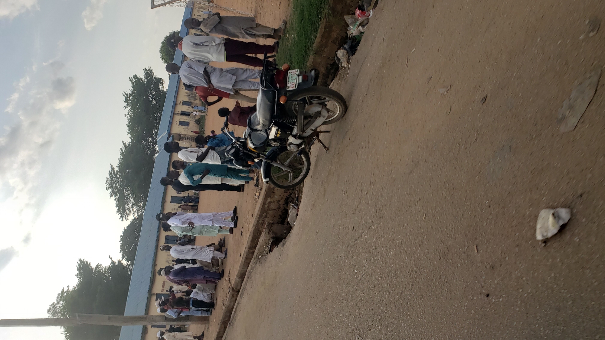 The image shows a street scene in Kawo, Nigeria. A group of people is lined up along a building with a blue roof. The people are wearing a variety of clothing. A motorcycle is parked on the edge of the road, near a curb. The road surface appears to be paved, and there is litter present. The sky is visible, with clouds. The image is rotated.