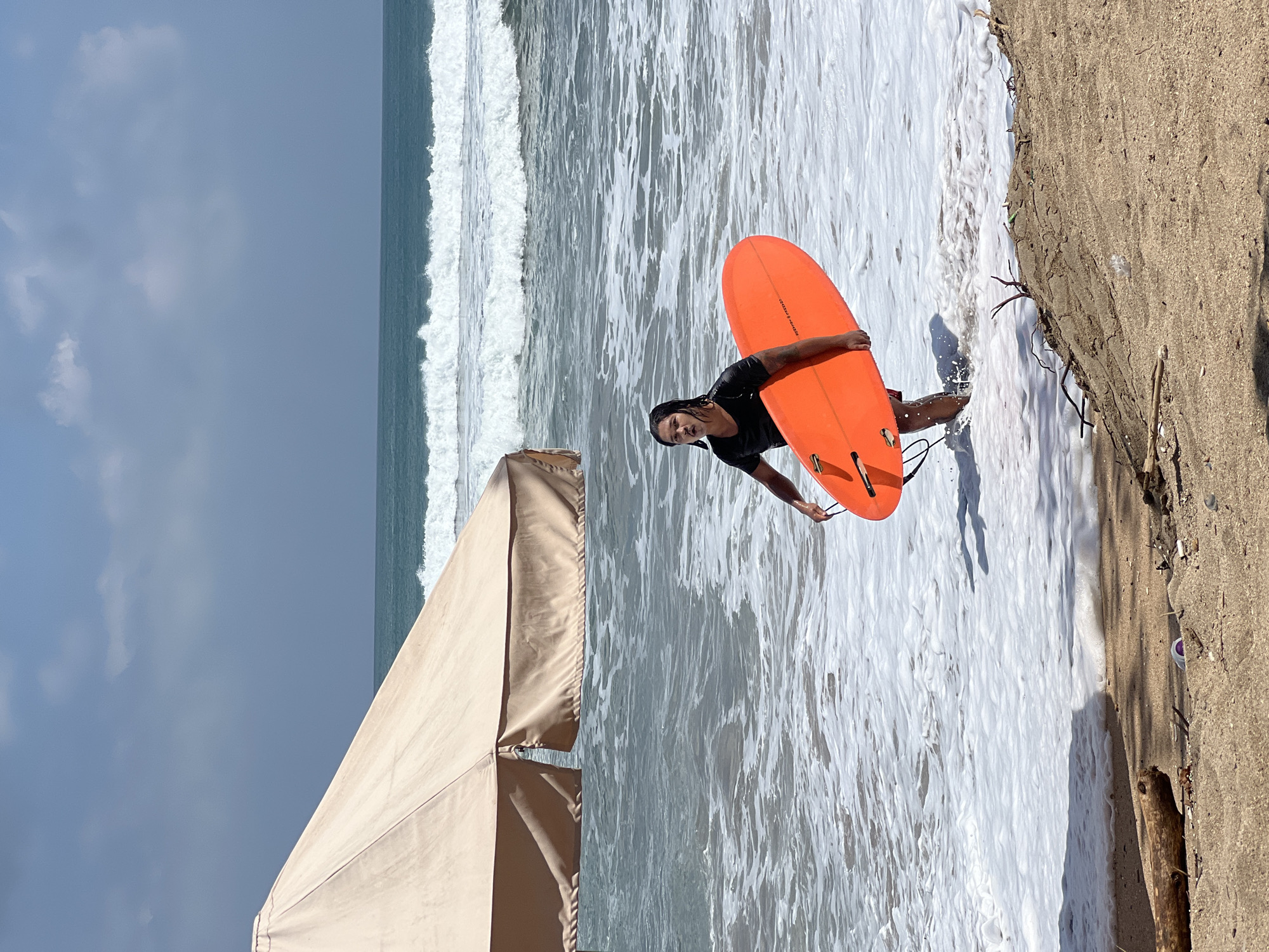 A person is walking out of the ocean in Kuta, Indonesia, carrying an orange surfboard. They are wearing a black t-shirt and shorts, and their hair is wet. The ocean waves are breaking behind them, and the sky is blue with some clouds. A large, light-colored umbrella is visible to the left of the frame, providing shade on the sandy beach. Debris, including a piece of driftwood and some plastic, is scattered on the sand in the foreground. The scene depicts a typical day at the beach, with recreational activity and natural elements.