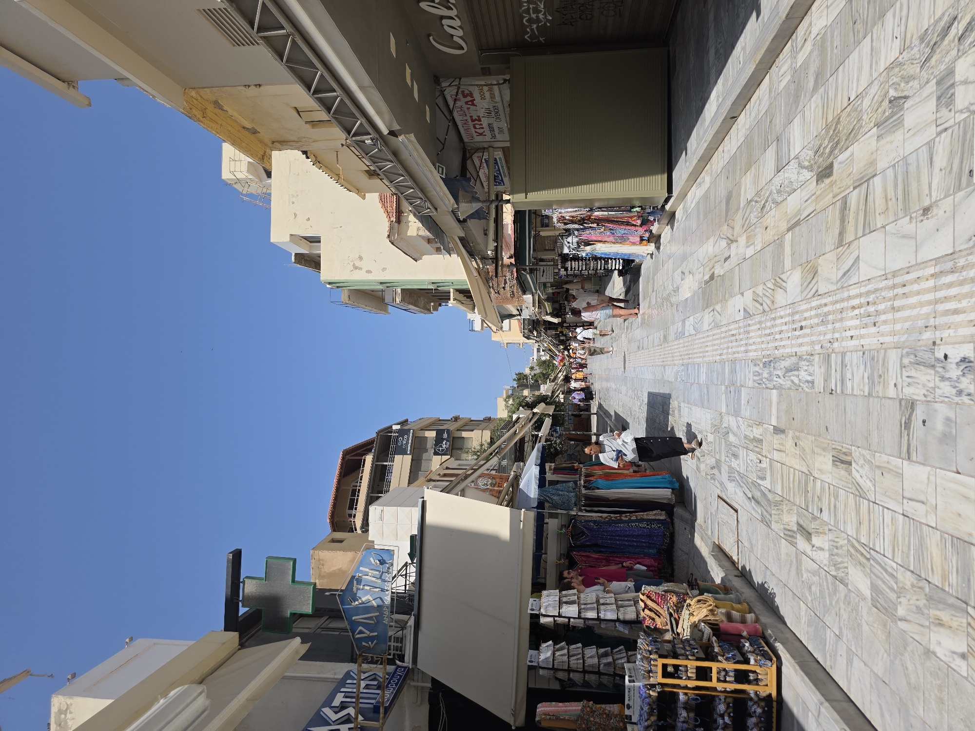 The image shows a street scene in Heraklion Municipal Unit, Greece, with shops lining both sides of a marble-paved pedestrian walkway. The day is bright and sunny with a clear blue sky.
On the left side of the street, shops display clothing on racks and shelves. A prominent green cross sign, commonly associated with pharmacies, is visible above one of the shops. The shop awnings provide shade over the displays.
On the right side of the street, similar shops are present, some with their shutters down. One shop displays signage with Greek lettering and brand names like "Calo" and "ROCHAS." Graffiti is visible on a shuttered storefront.
Several people are walking along the street. A woman in a white top and black skirt is walking towards the viewer on the left side of the walkway. Other pedestrians are seen further down the street, appearing to browse or move between shops. The marble paving stones are laid in a rectangular pattern, with a tactile paving strip running along the left edge of the walkway. The overall impression is of a bustling commercial street in a Mediterranean city.