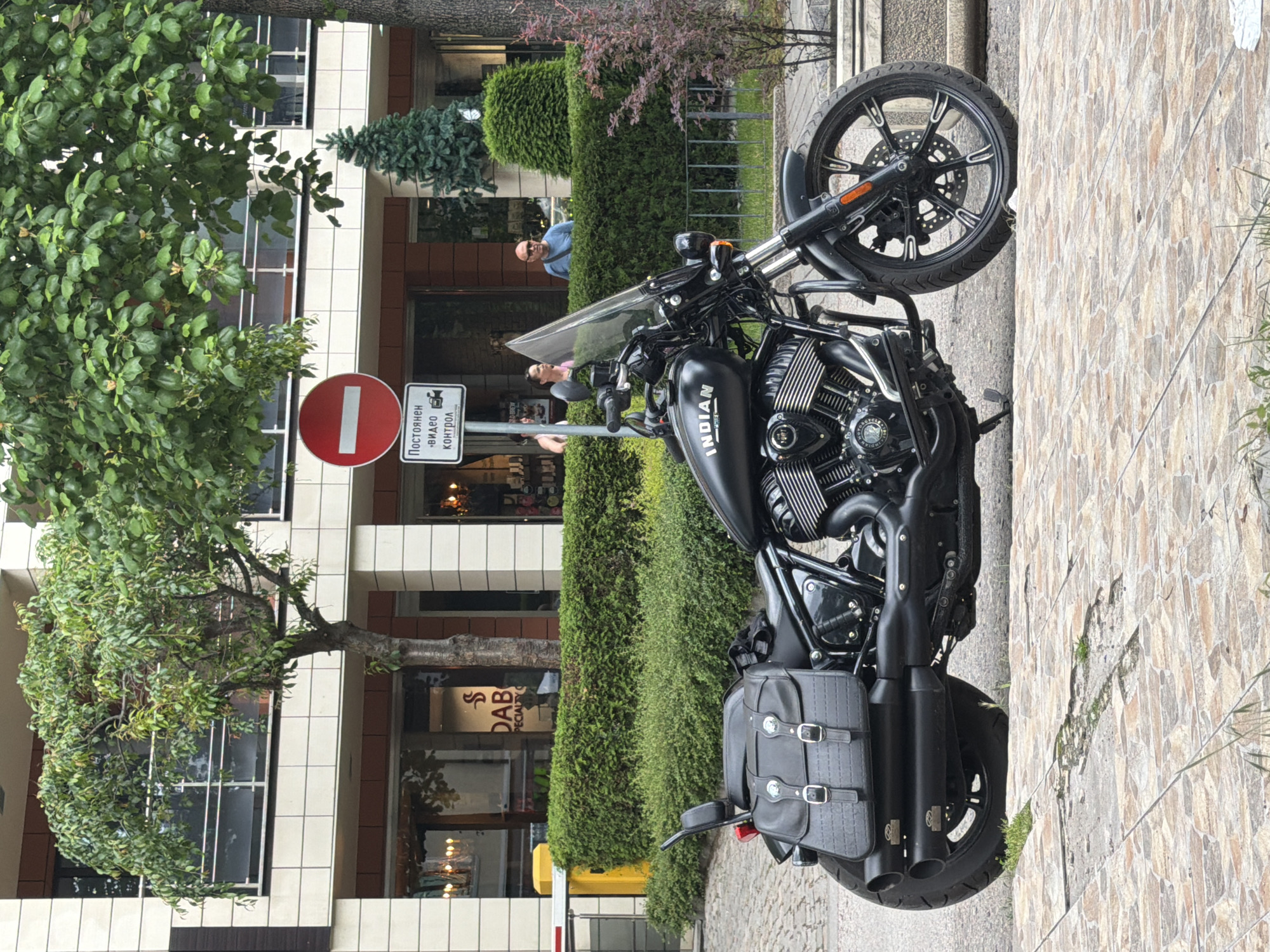 A black "Indian" brand motorcycle is prominently parked on a street in Sofia, Bulgaria, captured from a slightly elevated side angle. The motorcycle features a clear windshield, a detailed engine, and black leather saddlebags mounted on the rear.
Behind the motorcycle, a modern multi-story building with white and dark-paneled facades rises, partially obscured by vibrant green trees and neatly trimmed hedges. Two individuals are partially visible in the background: a man in sunglasses and a blue shirt peers over a hedge, while another person in a pink top is seen behind the motorcycle's windshield. A third person's arm is also visible further to the left.
To the left of the motorcycle, a red circular traffic sign with a white horizontal bar (indicating "No Entry" or "Do Not Enter") is mounted on a pole. Below it is a white rectangular sign displaying a camera icon and the Cyrillic text "Постоянен видео контрол," which translates to "Permanent video control."
The setting appears to be a city street or sidewalk, paved with textured, light-colored tiles on the right and a grey, paved surface beneath the motorcycle. The time of day is daytime, with soft, diffused lighting suggesting an overcast sky or early/late hours. The overall scene depicts a typical urban environment.