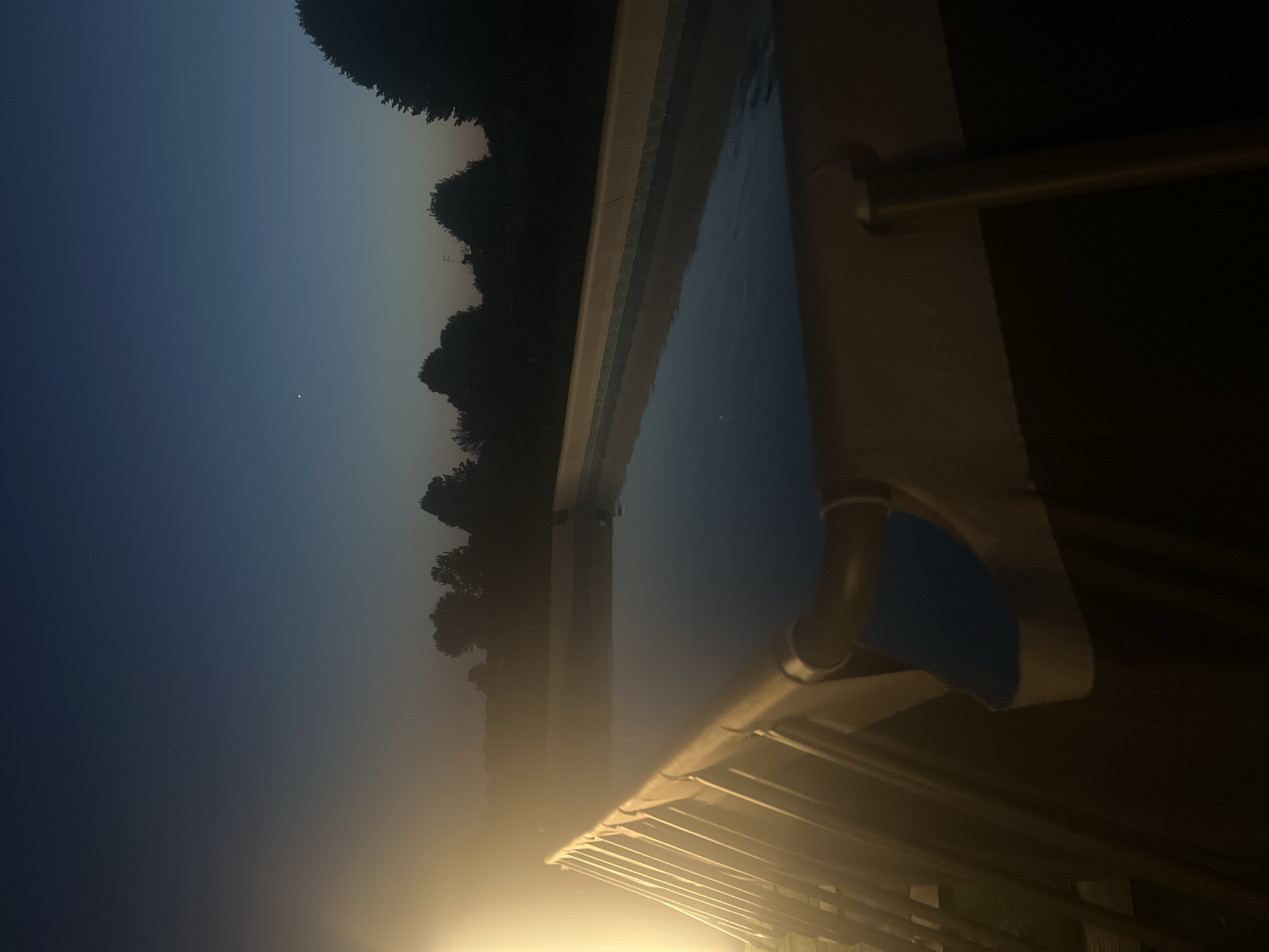 The image shows an outdoor above-ground swimming pool at dusk in Arre, Italy. A bright light source, possibly artificial, illuminates the left side of the pool and the surrounding area, creating a hazy glow. The sky above is a gradient of deep blue, transitioning to a lighter blue near the horizon, with a single star visible. Silhouetted trees form the distant treeline. The pool itself is rectangular, filled with dark water that reflects the dim light of the sky. The metal frame of the pool is visible, along with a blue liner section. The overall scene is peaceful and serene, capturing a moment as day transitions into night.