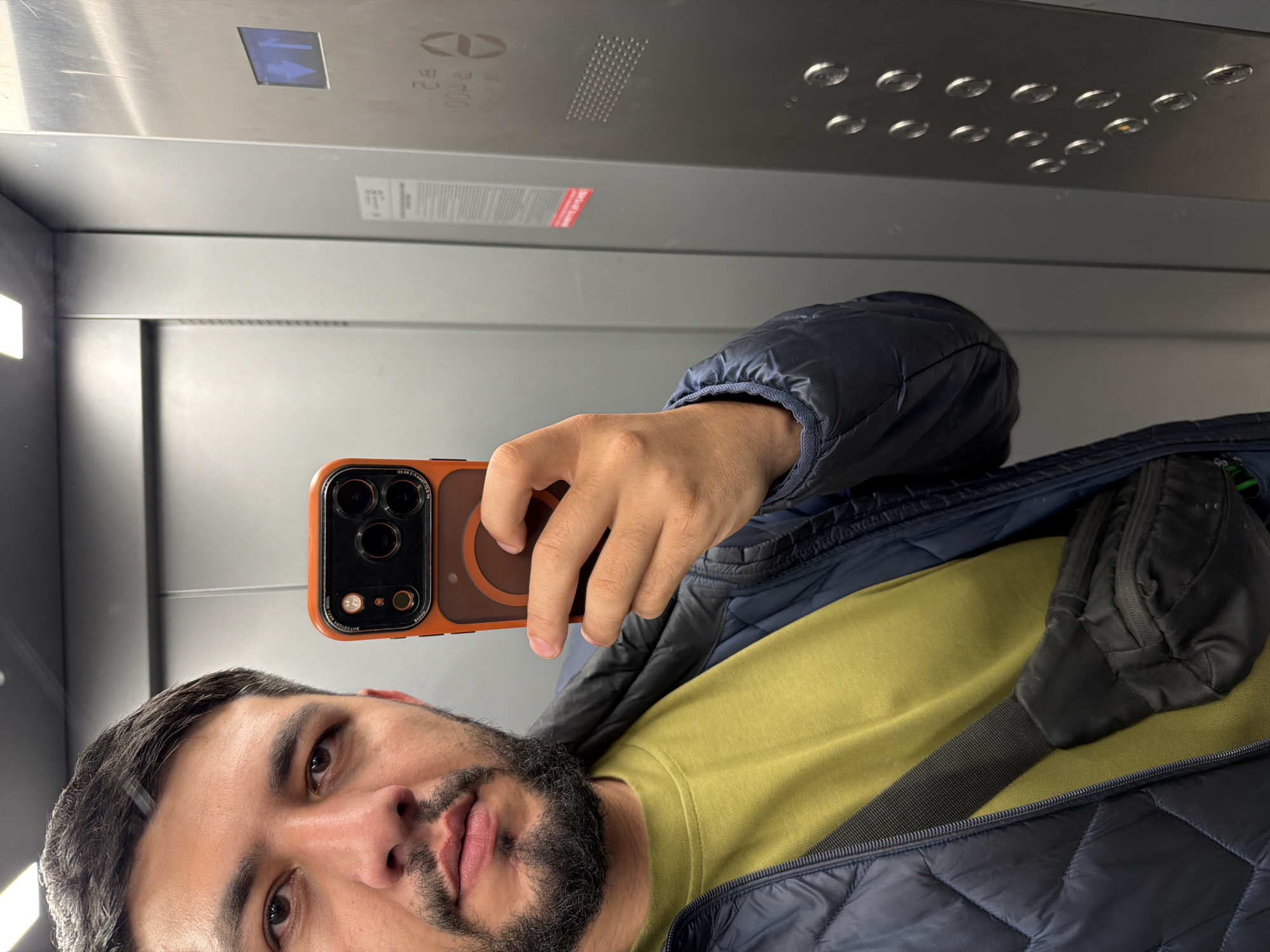 A man is taking a selfie in an elevator. He is holding a phone with an orange case. He is wearing a green shirt under a blue quilted jacket, with a black fanny pack slung across his chest. The elevator has a metal interior with a panel of buttons and an arrow pointing upwards. The elevator is identified as produced by Electrogorsk, a Russian manufacturer. The image was likely taken in Krasnoyarsk, Russia based on the manufacturer of the elevator and the man's appearance.