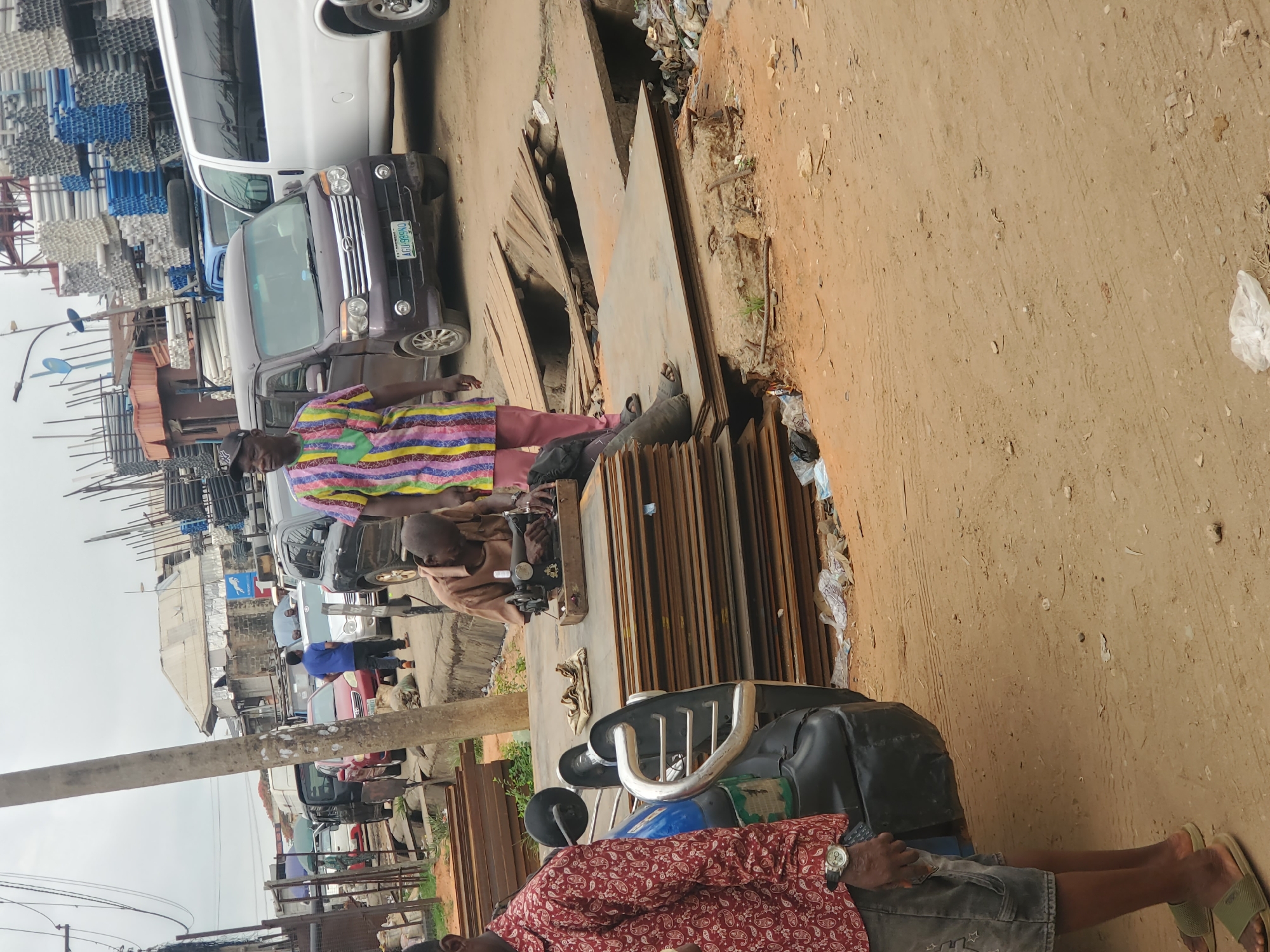 The image depicts a street scene in Fegge, Nigeria. A man in a colorful striped shirt and red pants stands near a stack of metal sheets on the side of a dirt road. Another man is sitting and appears to be operating a sewing machine on top of the metal sheets. A motorcycle is parked in front of the metal. The foreground features the legs of a man wearing a red patterned shirt, grey shorts, and sandals. Several vehicles, including a white van and a purple car, are parked along the street in the background. Stacks of metal pipes and buildings are visible in the distance. The ground is dirt and there is some trash scattered around.