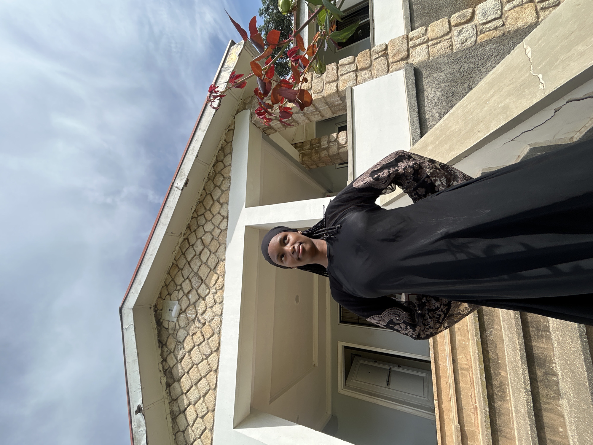 A young woman, wearing a black hijab and a black abaya with intricate lace detailing on the sleeves, stands on the stairs of a building. The building has a stone facade and a white overhang. Red leaves from a tree are visible in the upper left corner of the image. The sky is partly cloudy. The image appears to have been taken during the daytime. The woman is looking up and to the left, with a slight smile on her face. The overall scene suggests a pleasant, possibly casual, outdoor setting. The location context indicates Vom, Nigeria.