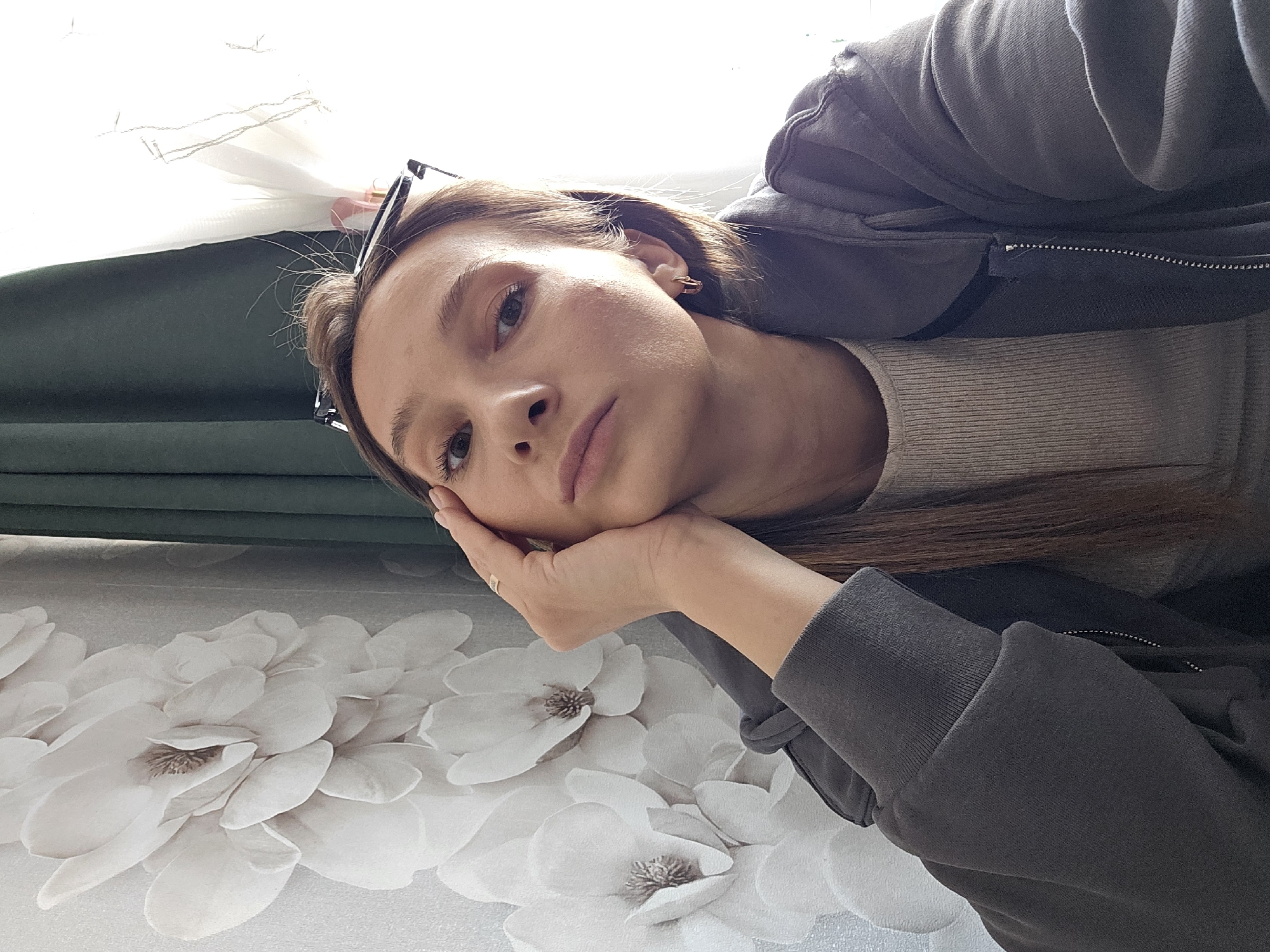 The image captures a young adult female indoors, located in the city of Chaikovsky, Russia. She is depicted from the chest up, facing forward with a direct gaze toward the camera, her right hand resting with the palm against the right side of her face and jawline. She has light brown hair, with some pulled back and dark-framed glasses positioned on top of her head. She wears a light-colored, possibly beige or light grey, ribbed crew-neck top beneath an open dark grey zip-up hooded sweatshirt. A simple gold-toned band ring is visible on the ring finger of her right hand, and small hoop earrings are present in her ears. The background to her left consists of a wall covered in light grey wallpaper patterned with large, white, multi-petaled flowers, each with a darker center. To her right, a window area is visible, partially obscured by a dark green curtain on the left and a brightly lit sheer white curtain or blind on the right, admitting significant natural light.