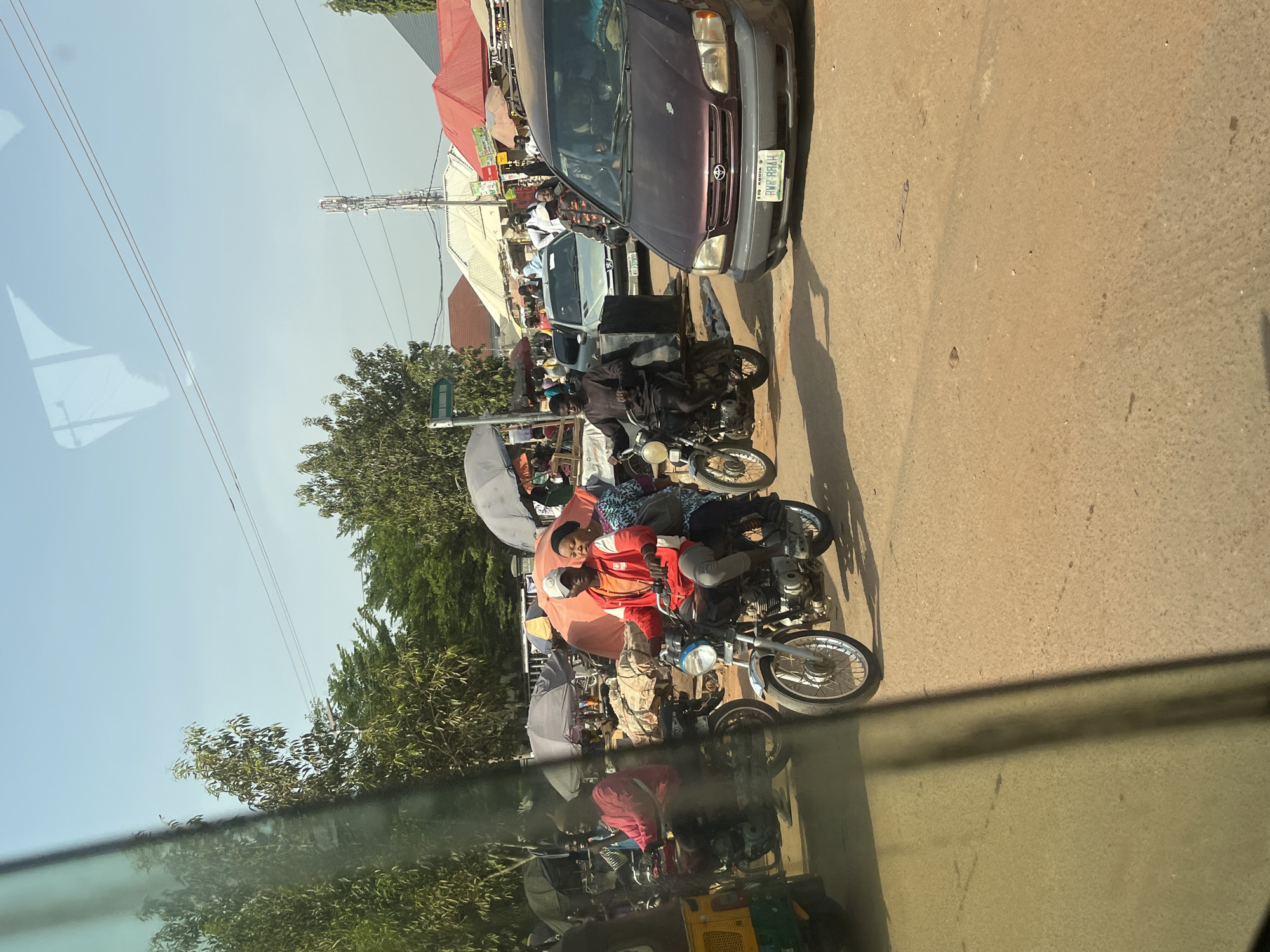 This media file captures a bright, sunny daytime scene of a busy street, likely in Nigeria, as indicated by a vehicle's license plate. The image appears to be taken from inside a vehicle, looking out, evidenced by the reflection of trees and other elements visible in the lower foreground.
The street is populated with various vehicles and people. A dark purple Toyota car is prominently featured on the right side of the road, facing towards the right. Its license plate is clearly visible, reading "ADAMAWA" (a state in Nigeria) and "FGH-884AH". Behind this car, another lighter-colored vehicle can be seen.
Several motorcycles are present, some carrying passengers. In the mid-ground, a person wearing a red top and a light-colored cap is seated on a motorcycle, with another person behind them. They are positioned next to an orange umbrella, suggesting a roadside stall or waiting area. Further down the street, another individual on a motorcycle is visible in a dark outfit.
Along the left side of the road, there are market stalls or vendors, many using umbrellas (white and orange/red) for shade. Green trees and foliage line this side of the street. The sky is clear and blue, with power lines running across it. In the distance on the right, a communication tower or a tall utility pole stands against the sky. The road surface appears to be asphalt or packed earth, with some debris. The strong sunlight casts clear shadows, indicating midday or early afternoon.