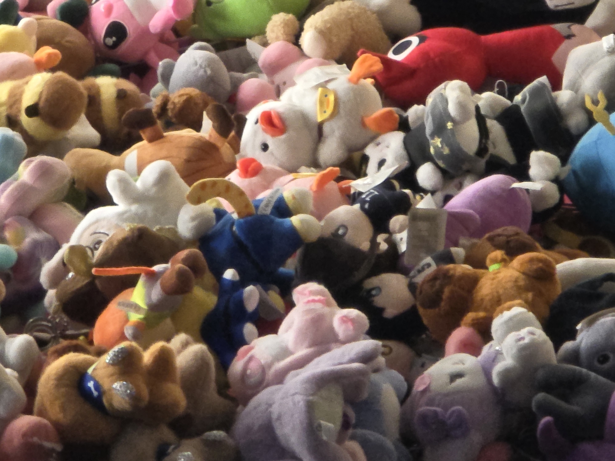 The image displays a close-up, vertical view of a large collection of stuffed animals. The stuffed animals are densely packed and fill the entire frame, creating a colorful and chaotic arrangement. Dominant colors include various shades of brown, pink, purple, blue, white, and green. Several stuffed animals are visible in detail, such as a white duck-like creature with orange feet and beak wearing a yellow bib, a pink creature with large eyes, and a red, elongated creature with black eyes. Other stuffed animals appear to be dogs, cats, and various cartoon characters. The lighting suggests an indoor environment, possibly a store or a display. There are no people visible in the image, and the stuffed animals are stationary. The overall scene is one of abundance and variety in plush toys.