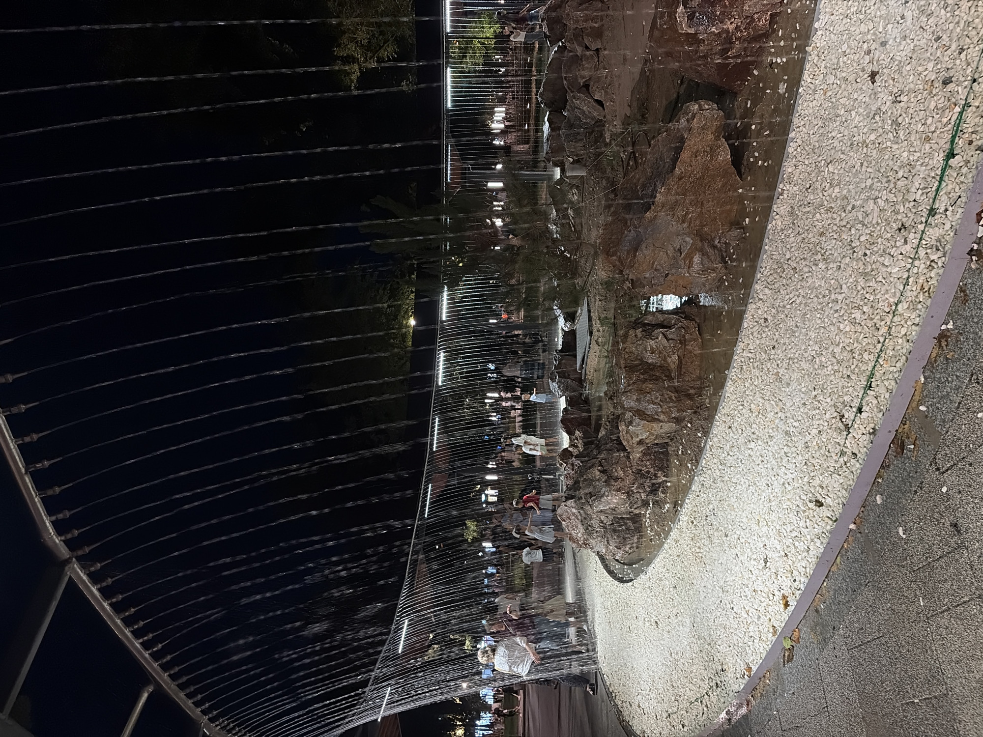 The image depicts an outdoor water feature at night, possibly located in the city of Shymkent, Kazakhstan. A curved structure above emits streams of water that fall into a rocky basin filled with water. People are visible in the background, some interacting with the water feature. The ground surrounding the basin is covered in white gravel. The scene is illuminated by artificial lighting.