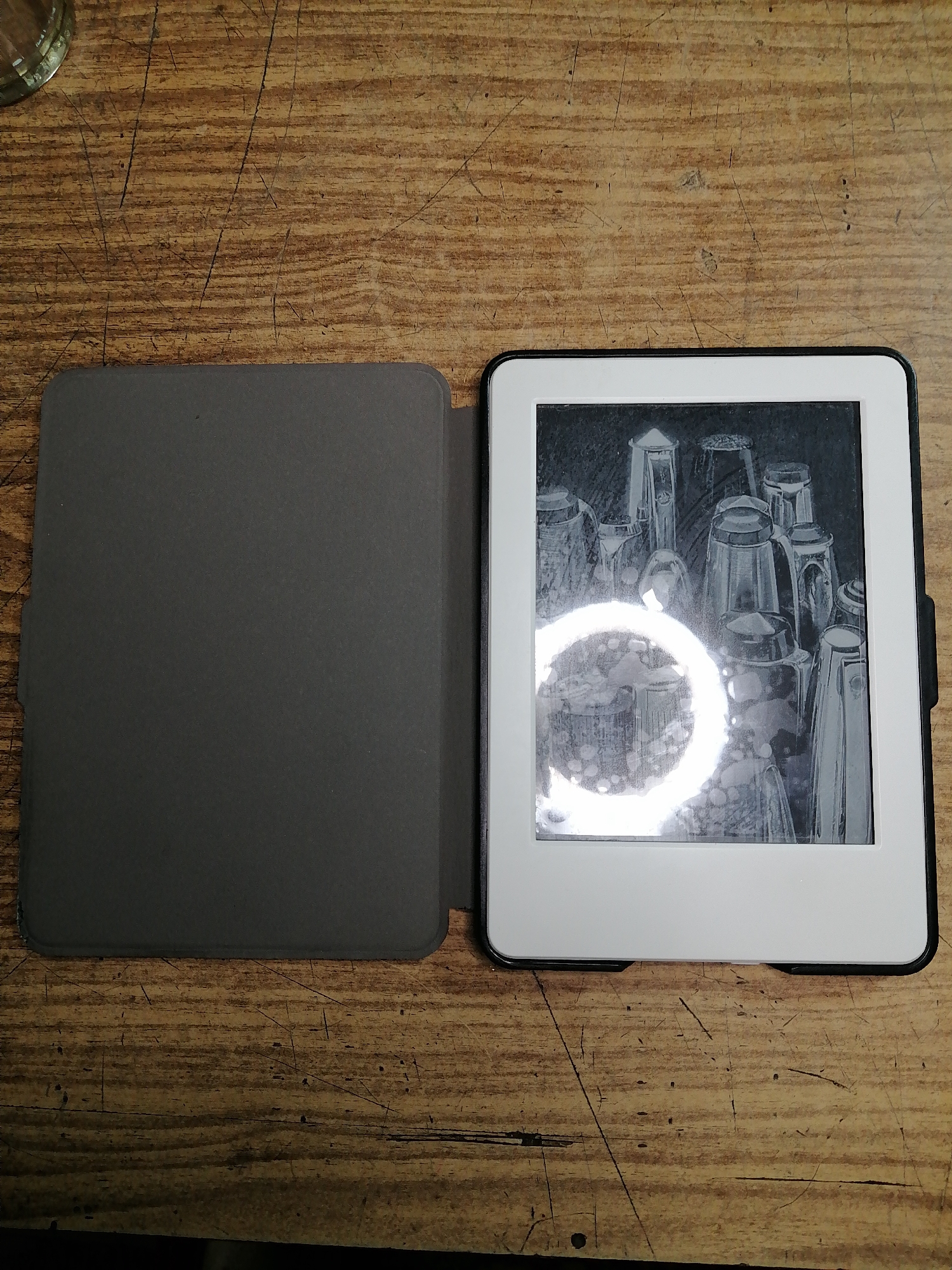 The image shows an e-reader with a dark gray cover open on a wooden surface. The e-reader screen displays a black and white image of a collection of glasses. The wooden surface appears worn with visible grain and scratches. The overall scene is lit by overhead light, creating a bright circular reflection on the e-reader screen. There are no people or animals visible in the image. The location of this scene is not determinable from the visual information provided.