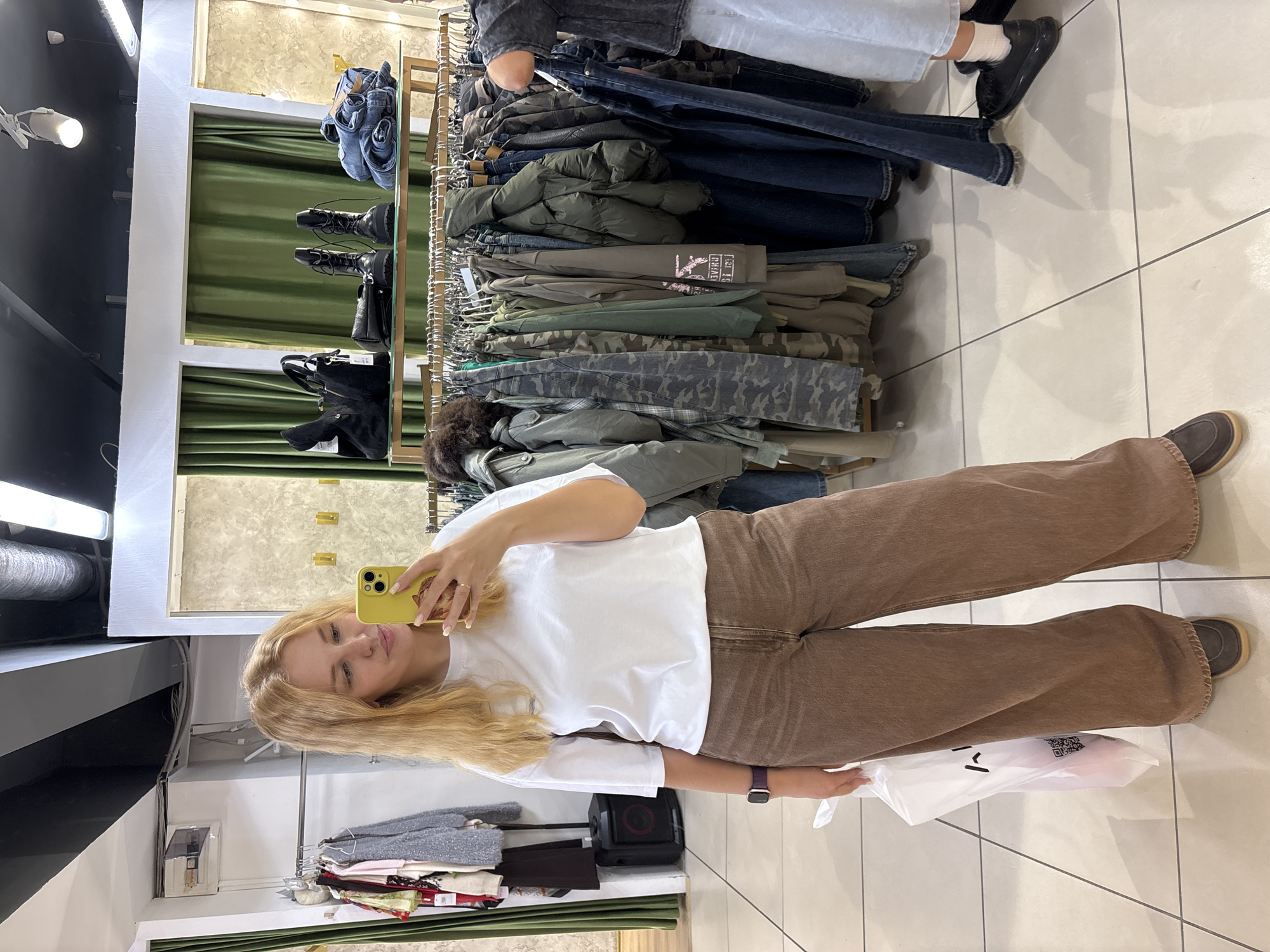 The image depicts a young woman taking a mirror selfie in a clothing store. She has blonde hair and is wearing a white t-shirt and brown wide-leg pants. She is holding a white shopping bag in her left hand and her phone in her right hand. In the background, there are racks of clothing, including jackets, shirts, and jeans, as well as a display shelf with handbags and boots. The store appears to be well-lit with fluorescent lighting and has a tiled floor. The location is identified as the city of Rusanovo, Russia.