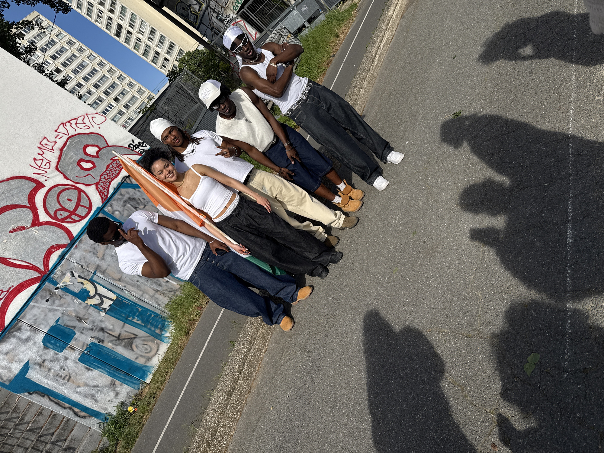 Six young individuals are posing diagonally across a paved outdoor area under bright sunlight in Charenton-le-Pont, France. The group consists of four males and two females, all appearing to be of Black ethnicity, dressed in casual streetwear. Their attire predominantly features white tops, baggy jeans or shorts, and accessories such as hats, sunglasses, and chains. Many are smiling or striking poses for the camera.
To their left, a white wall is extensively covered in red and grey graffiti, depicting a stylized skull or face and various abstract patterns. Words like "VIEJO" appear to be part of the graffiti. Behind this wall, tall, modern multi-story residential buildings rise against a clear blue sky. To the right of the subjects, there's a paved road or path with a curb, patches of grass, and a metal fence.
One woman in the center of the group is holding an orange and white flag. Strong, elongated shadows are cast on the pavement, indicating a sunny day, likely morning or late afternoon. The overall atmosphere is cheerful and relaxed, capturing a moment of social interaction in an urban setting.
