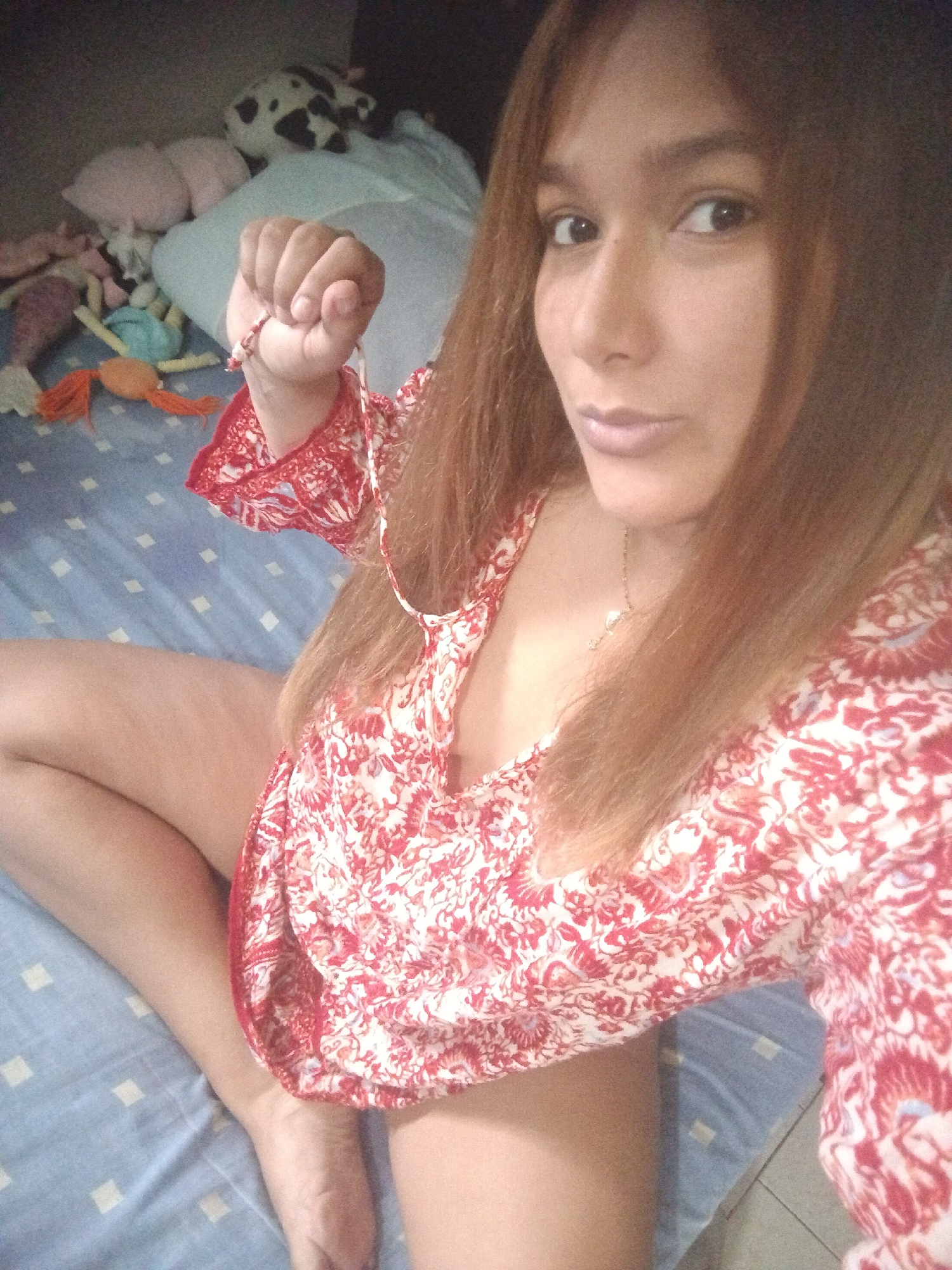 A person with brown hair and a red and white patterned top is sitting on a blue and white checkered bedsheet. They are looking at the camera with a slight pout. Their right fist is clenched, holding a small beaded bracelet. In the background, stuffed animals are visible. There is no contextual information available in the image to identify a specific location in Caracas, Venezuela.