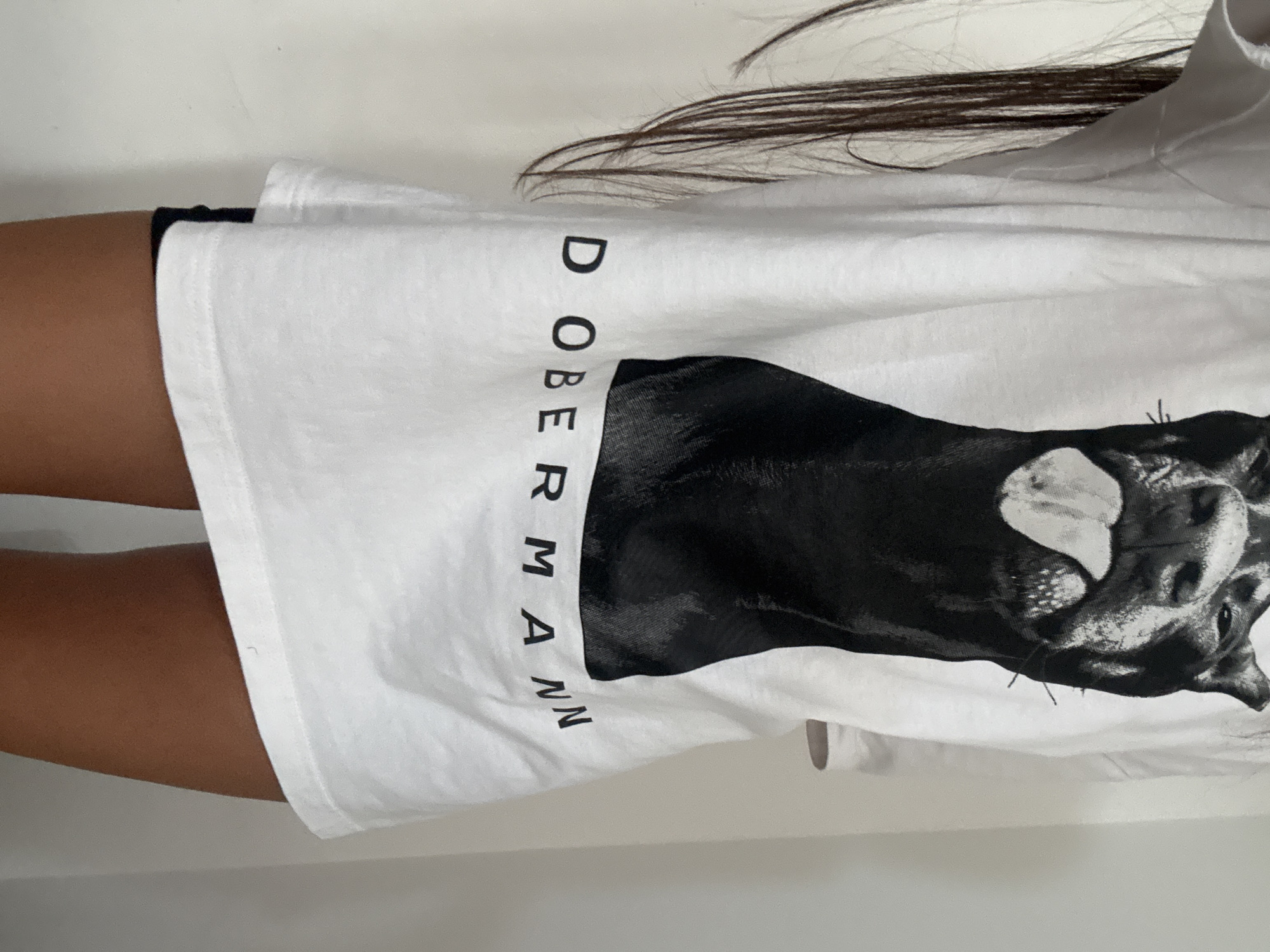A person wearing a white t-shirt with a large black and white image of a Doberman pinscher's head and the word "DOBERMANN" printed below it is shown from the waist down. The person's long dark hair is visible on the left side of the image. They are wearing black shorts. The background is a plain white wall. The image does not contain any elements that would allow for determining its location in Chișinău, Moldova.