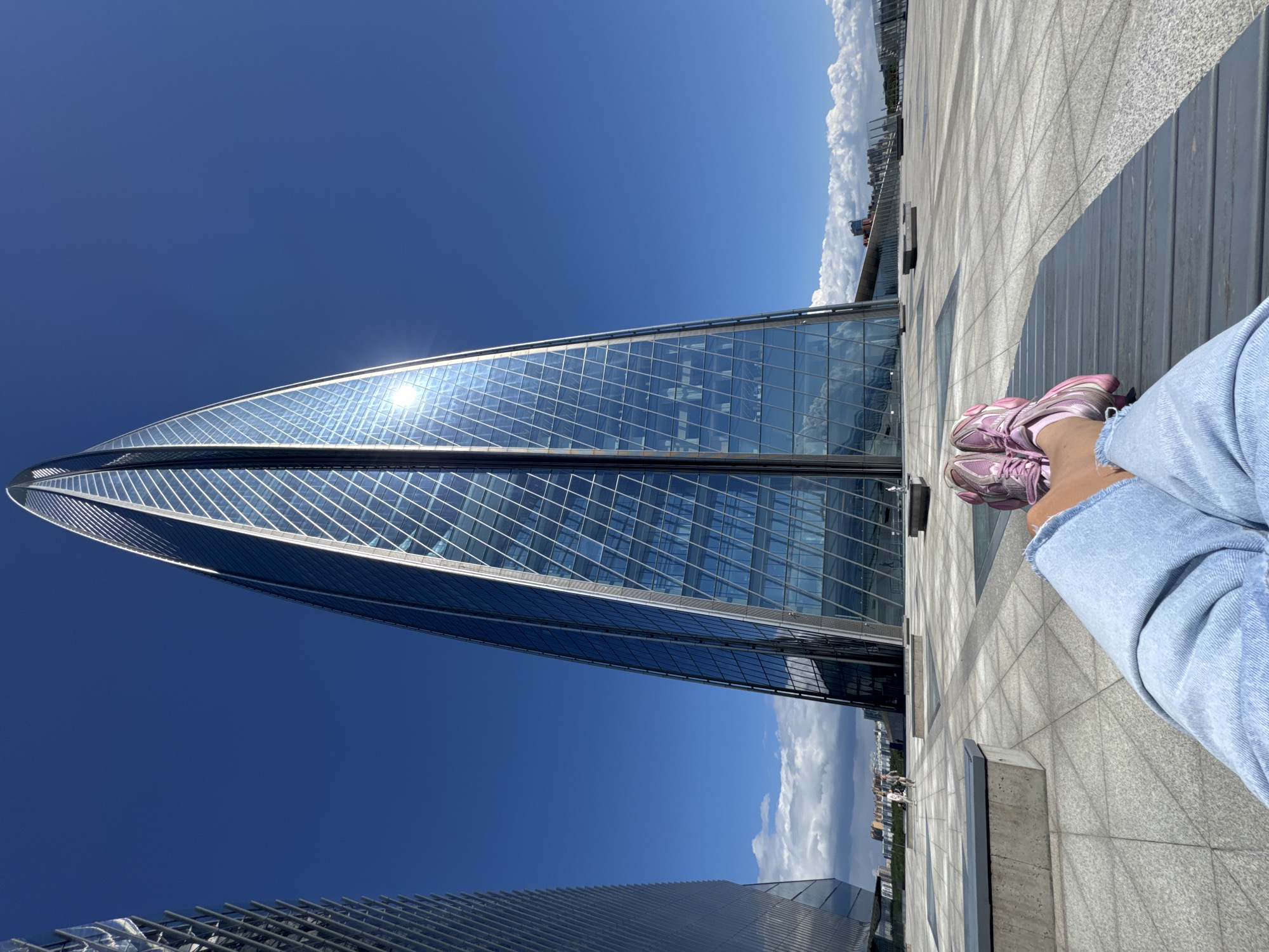 The image captures a vibrant, sunlit outdoor urban scene, featuring a person's lower body in the foreground and a striking modern skyscraper. The person, seen from the mid-thigh down on the right side of the frame, is wearing light blue jeans with a distressed hem and pink athletic sneakers. They appear to be sitting or resting on a light-colored wooden bench or surface.
Dominating the left side of the frame is a tall, slender, and distinctly twisted glass skyscraper. Its facade, composed of numerous reflective glass panels, brightly reflects the sun, creating a strong glare near its upper section. The skyscraper's unique architectural design suggests a modern landmark.
The setting appears to be an open plaza or rooftop terrace, paved with large, light-colored square tiles. The sky is a brilliant, clear blue, with scattered white clouds visible along the horizon and reflected in the glass surfaces of the buildings. The overall impression is one of a bright, clear day in a contemporary city. This scene is located in Saint Petersburg, Russia. There is no visible text in the image.