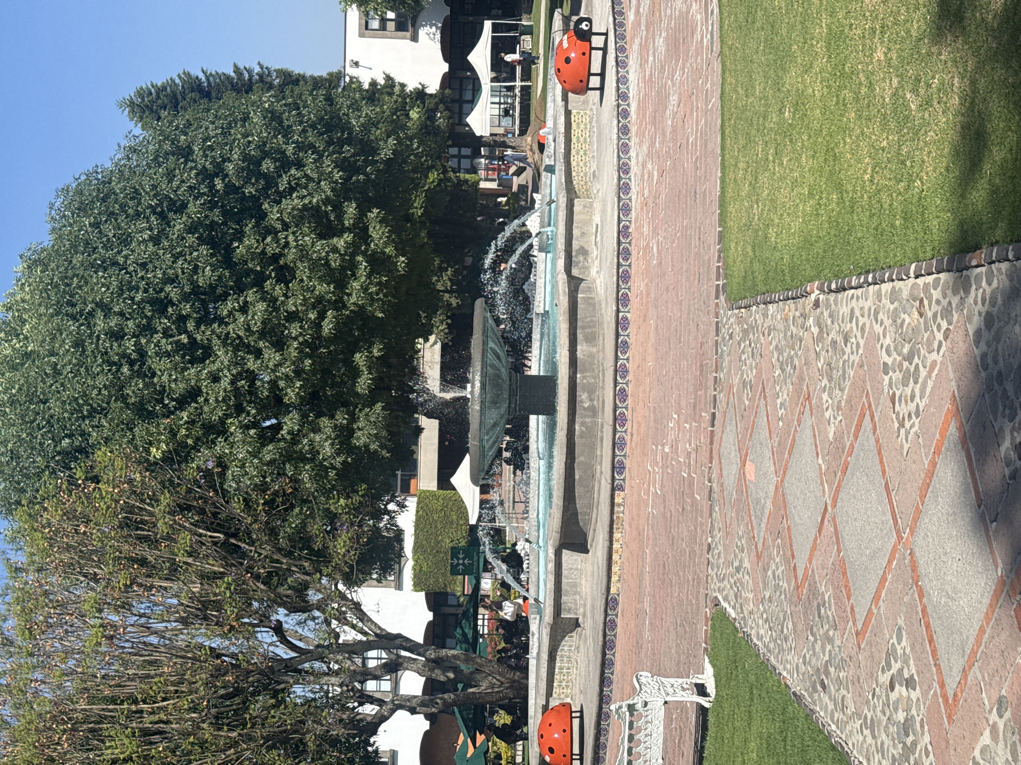 This outdoor scene, likely in the city of San Andrés Cholula, Mexico, shows a park-like area on a clear day. A central fountain with multiple tiers of water is the focal point. Red and black ladybug statues flank the edges of the fountain. Lush green trees surround the area, and white buildings are visible in the background. A brick-paved walkway borders a section of green grass, with diamond-patterned paving and pebbles visible. People are present but appear small and indistinct due to the distance.