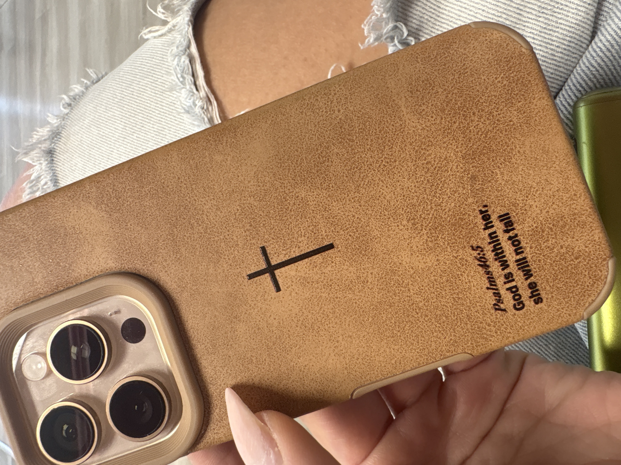 A person is holding a tan phone case with a Christian cross and the text "Psalms 46:5 God is within her, she will not fall." The phone case has a camera unit with three lenses. The person's hand and part of their nails are visible. They are wearing a denim shirt or jacket. The background includes a striped shirt or jacket. The location is unknown and cannot be determined to be Plovdiv, Bulgaria.