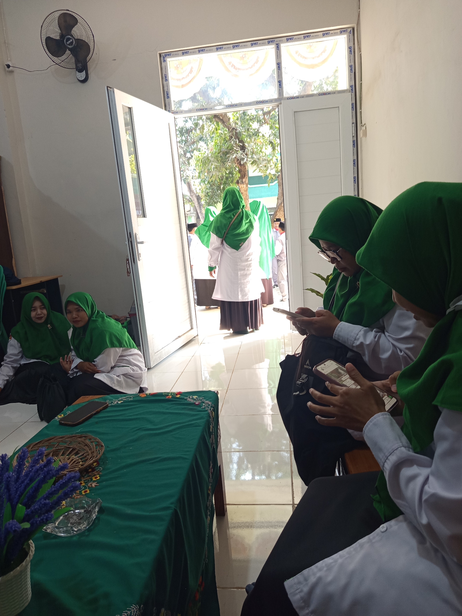 In Lumutan, Indonesia, an indoor gathering is captured. The scene depicts a group of young women, primarily dressed in green hijabs and white shirts. Some are seated on the floor, while others are standing. A woman in the background, also wearing a green hijab and white attire, is walking through an open doorway. Two women in the foreground are engrossed in their mobile phones. A table draped with a green cloth occupies the lower part of the frame, adorned with a woven basket, a clear glass ashtray, and a decorative plant with purple flowers. A ceiling fan is visible on the upper left wall, and a window with an aluminum frame looks out onto a leafy outdoor area. The floor is tiled. The lighting suggests a bright, daytime setting.