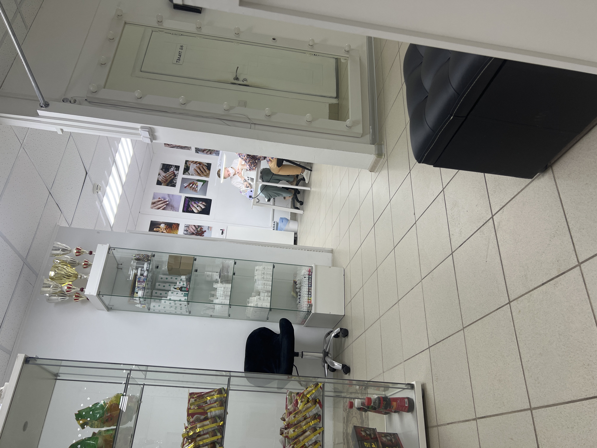 This image captures the interior of a brightly lit nail salon or beauty studio, likely in Chaikovsky, Russia, given the Cyrillic text visible. The perspective is from a slightly elevated position, looking into the main service area.
In the center, two individuals are engaged in a nail service. One person, presumably a nail technician, is seated at a white table, meticulously working on the hands of a client who is also seated opposite them. A UV lamp and other manicure tools are visible on the table.
The room is furnished with various items typical of such establishments. To the left, there are two glass display cases. The upper case holds numerous bottles of nail polish or beauty products, along with several golden trophies or awards on top. The lower display case contains packaged goods, possibly snacks or additional beauty products. A black office chair on wheels is positioned between the two cases.
Against the back wall, a prominent feature is a white door framed by a vanity mirror with light bulbs around it. On the door, reflected in the mirror, is the Russian word "ГЛАВНАЯ" (GLAVNAYA), meaning "MAIN". The wall next to the door is decorated with several framed pictures showcasing various manicured nail designs.
To the right foreground, a large, black, tufted ottoman or bench provides seating. The floor throughout the space consists of light-colored tiles with visible grout lines. The ceiling is a drop ceiling with integrated fluorescent lighting panels, illuminating the room evenly. The overall atmosphere appears clean and professional, with a focus on nail care services. No strong emotions are discernible from the individuals, and the time of day or external weather cannot be determined.