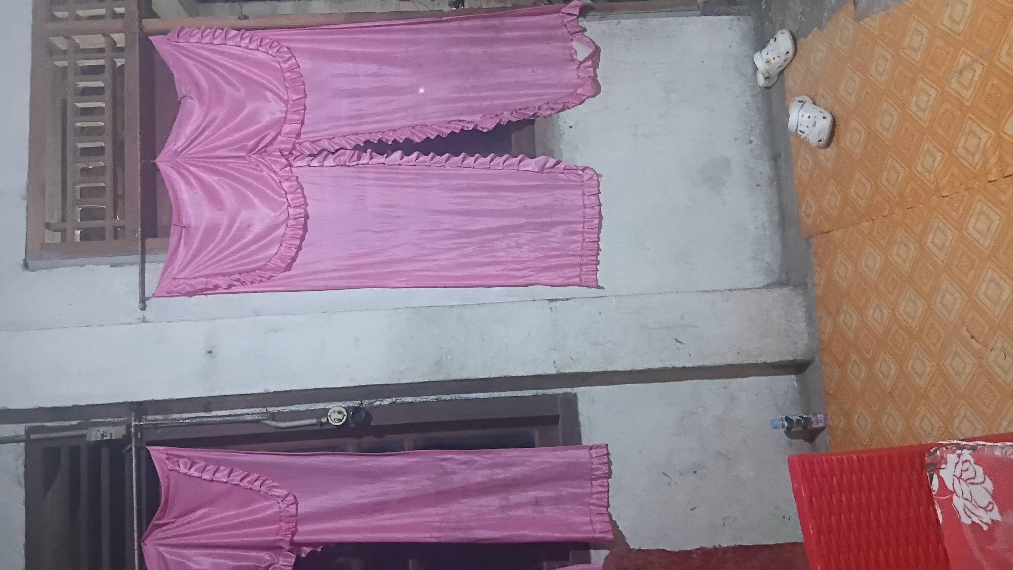 The image depicts an outdoor scene, likely a patio or veranda area, in front of a building with whitewashed walls and wooden window frames. Two sets of pink satin curtains with ruffled edges are hung over what appear to be windows or openings. One set of curtains is pulled to the left, revealing a dark interior, while the other set is partially open in the center. On the floor, which is covered with an orange and brown patterned mat, are two pairs of white slip-on shoes. To the left, a red woven chair is partially visible, and a small bottle of liquid stands near the wall. The overall impression is that of a domestic outdoor space. The image does not contain explicit information to pinpoint its location to the city of Prabumulih, Indonesia, or any specific city.