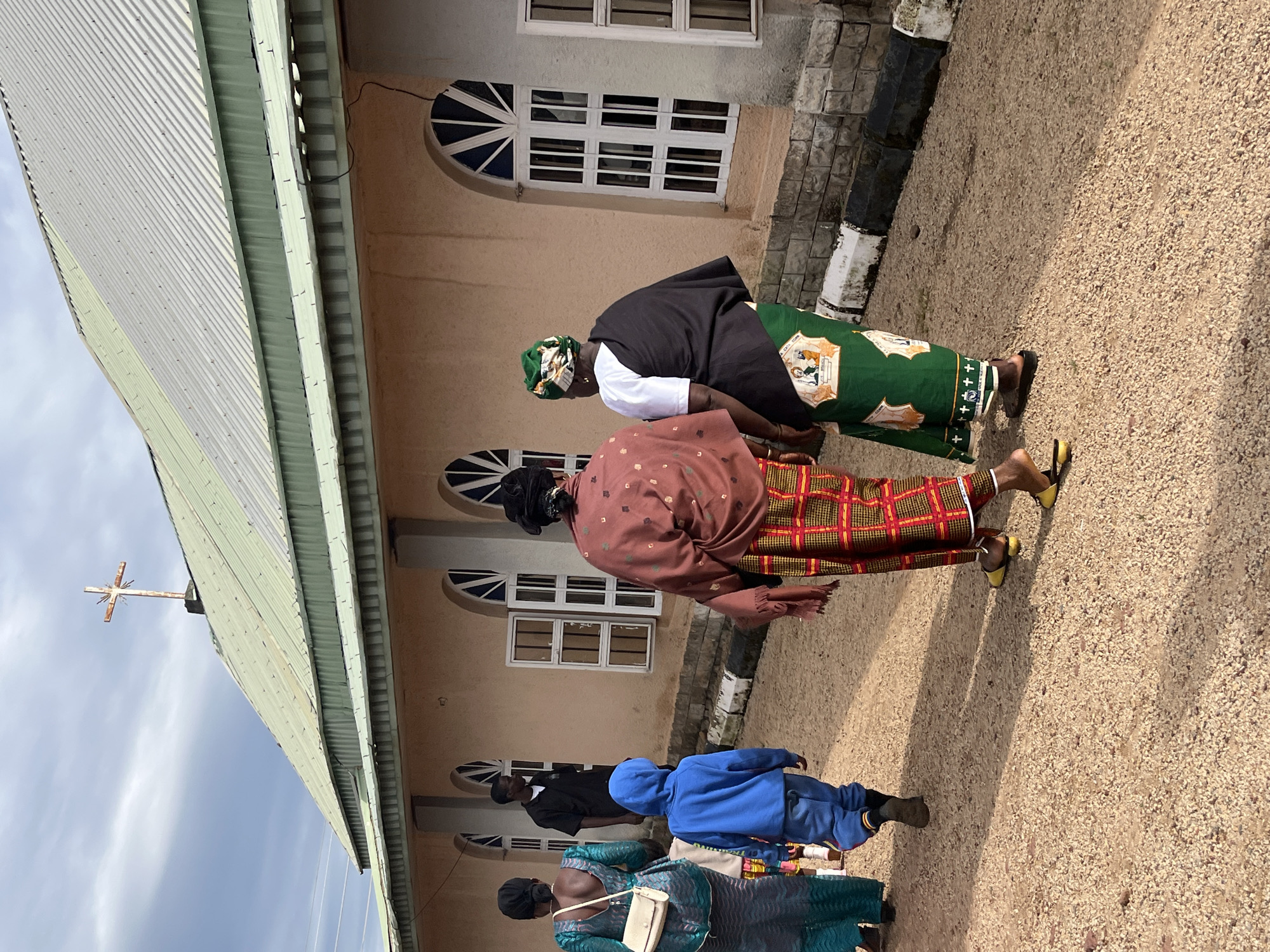 The image depicts a group of people walking away from the camera on a gravel path towards a building with a green-gabled roof topped by a rusty cross. The building appears to be a church, with arched windows and a beige facade. The people are dressed in a mix of traditional and modern clothing. Two women are in the foreground, one wearing a brown patterned wrapper and the other a green wrapper with an image of religious figures. A young boy in a blue hooded tracksuit walks in front of them, and a man in a black shirt stands behind them. The scene appears to be set in a semi-arid environment, possibly in Jos, Nigeria, given the architectural style and the clothing. The sky is cloudy, suggesting overcast weather. The lighting indicates it is daytime.
