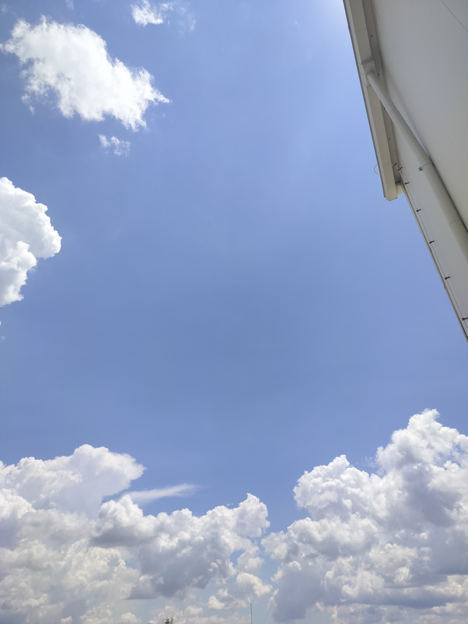 The image depicts a bright blue sky with scattered white cumulus clouds. In the upper right corner, a portion of a white building or structure with a gutter system is visible, angled downwards towards the sky. The lower part of the image is filled with more dense clouds, some appearing dark and heavy, suggesting potential for rain, while others are bright and fluffy. At the very bottom edge, what appear to be the tops of industrial structures or antennas can be faintly seen emerging from the clouds. The scene is a clear, daytime shot focusing on the sky and a partial view of an urban or industrial edge. There are no people or discernible actions visible.