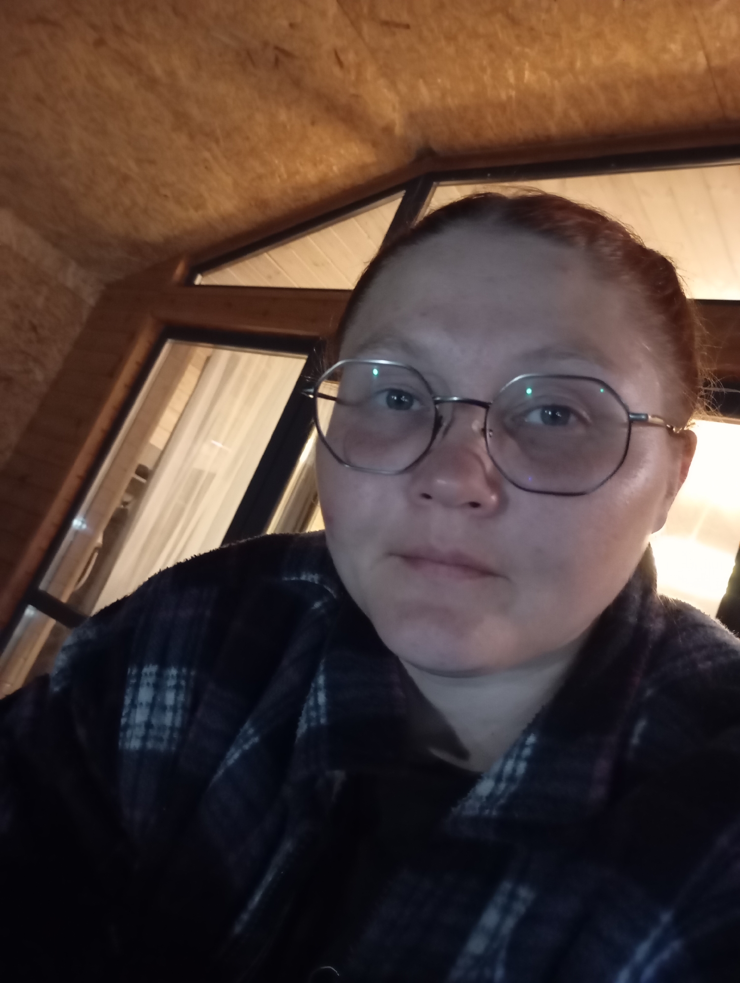 The image shows a close-up, slightly blurry selfie of a person with fair skin, wearing round, wire-rimmed glasses and a dark, plaid shirt. The person's hair appears to be pulled back. The background suggests an indoor setting with wooden walls and a window that lets in light, possibly from outside. The ambient lighting is warm and dim. The specific location in Malyi Bukor, Russia, is not discernible from the visual information provided in the image.