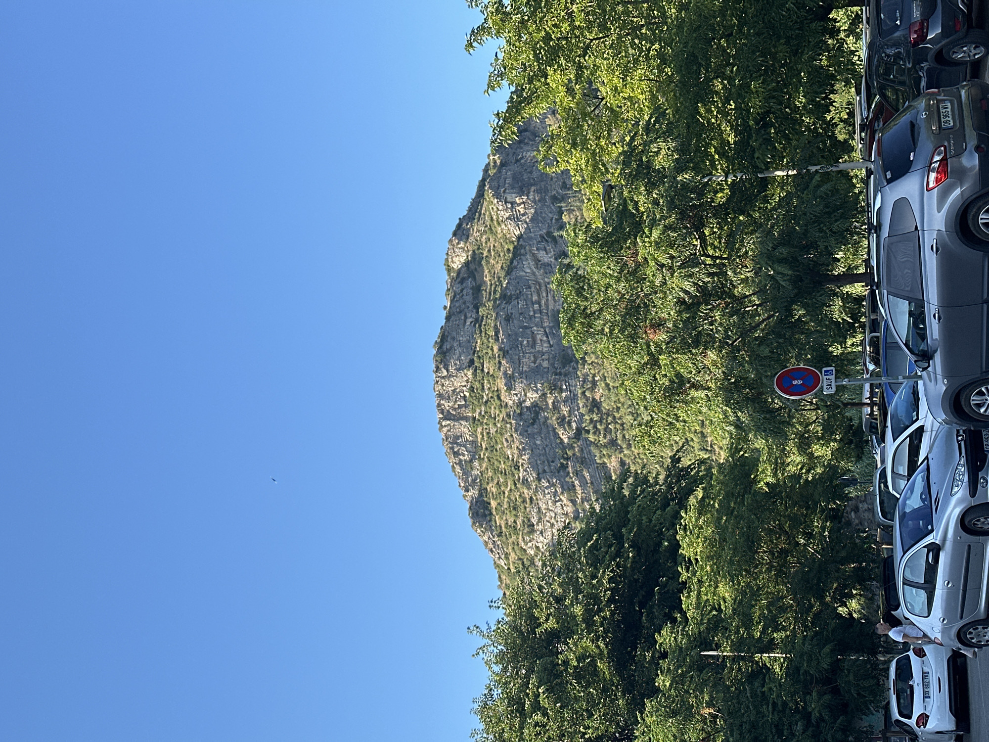 The image shows a steep, rocky mountain face partially covered in greenery, rising against a clear blue sky. In the foreground, a line of parked cars is visible, suggesting a roadside or parking area. Lush green trees with dense foliage are situated between the cars and the mountain, casting shadows and adding to the natural setting. The sun appears to be high in the sky, indicated by the bright daylight and the strong shadows.
One of the cars has a license plate visible that reads "CB-985-NV". There are also traffic signs, including one that appears to be a circular red and blue sign with a symbol in the middle, and another with the word "SAUF". A person in a white t-shirt is visible near one of the cars, standing on the right side of the image. The overall impression is a scenic view of a natural landscape with man-made elements like cars and signs present. The time of day is likely midday due to the lighting conditions.