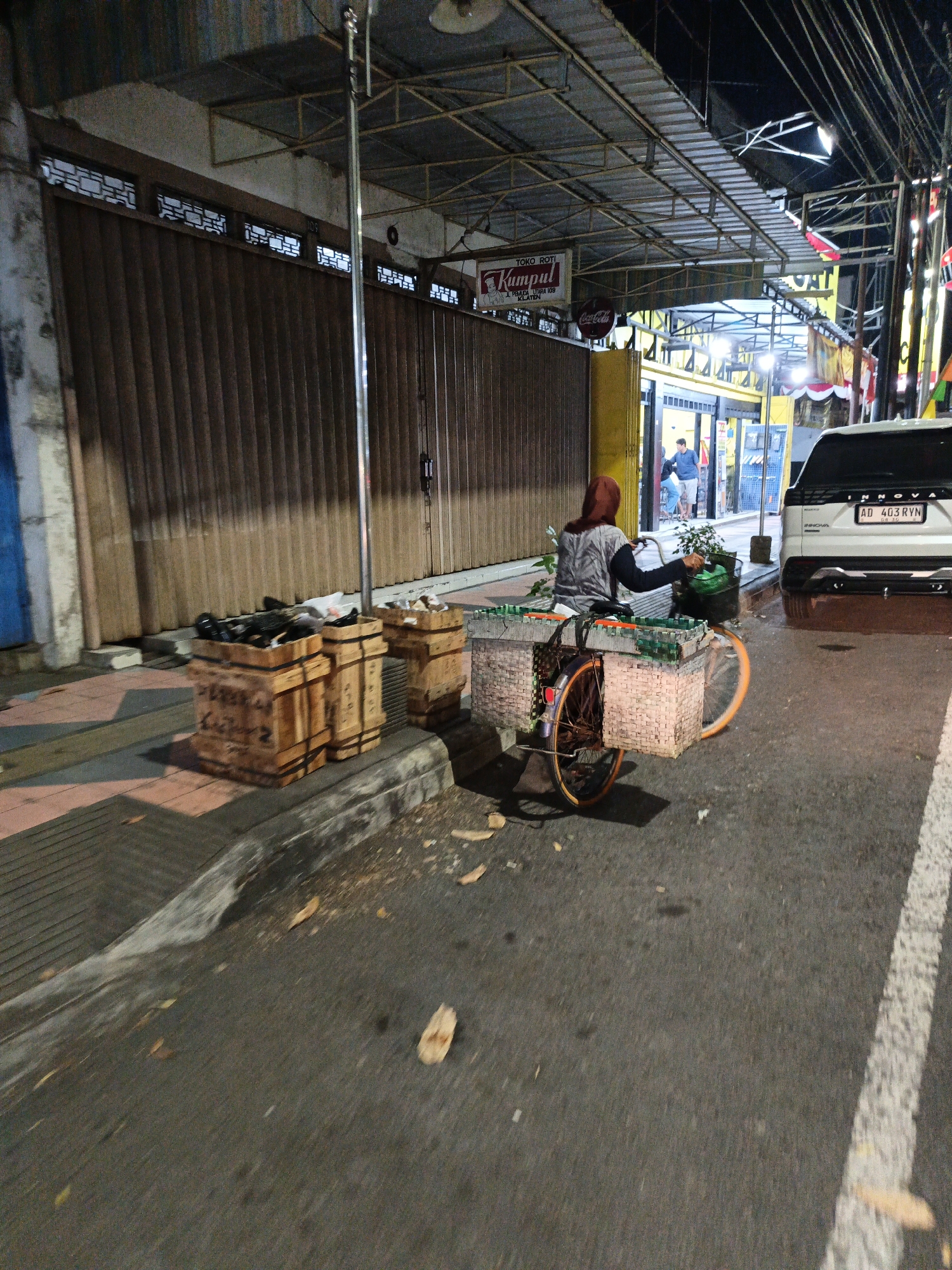 A person wearing a red hijab and a grey shirt is sitting on a bicycle at night on a city street in Klaten, Indonesia. The bicycle has two large woven baskets attached to the rear, one on each side, and a smaller basket on the handlebars. The person is holding a green bag in their right hand. The street is paved and has a white line marking the edge of the road. On the left side of the street, there are several wooden crates on the sidewalk. Behind the person and bicycle is a building with a large corrugated metal gate and a sign that reads "TOKO ROTI KUMPUL JL. PANGLIMA UTARA 109 KLATEN." A white SUV with license plate "AD 403 RYN" is parked on the right side of the street. The scene is lit by artificial light sources, suggesting it is evening or night.