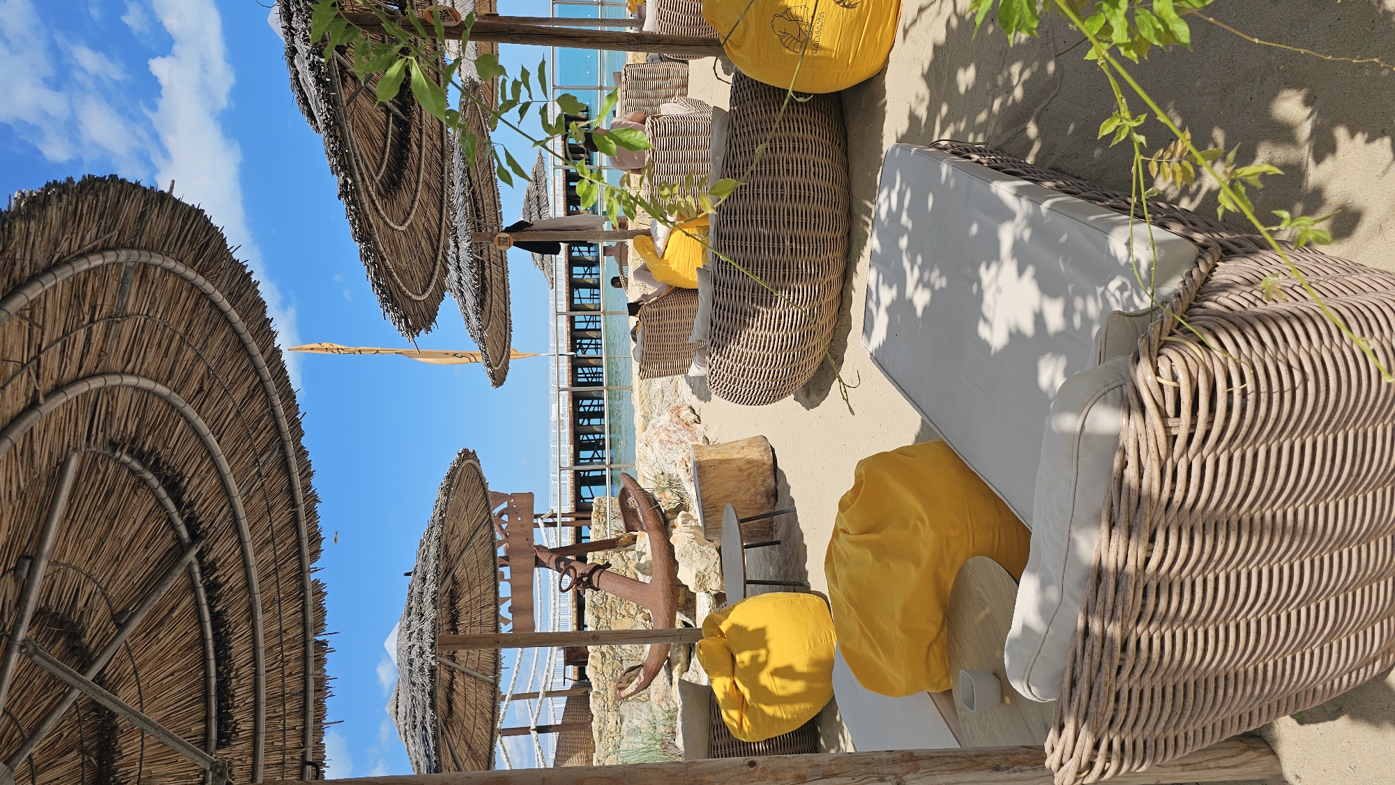 The image depicts an outdoor seating area on a sandy beach, likely a cafe or bar, near a pier extending into the sea. The area is furnished with woven wicker seating, including a long daybed in the foreground, and several round lounge chairs. Bright yellow beanbag chairs are scattered throughout the scene. In the background, a weathered anchor rests on a pile of rocks, with a wooden pier visible extending into the calm, blue water. Several thatched umbrellas provide shade. The sky is clear blue with a few wispy clouds. The overall ambiance suggests a relaxed, tropical setting. Based on the provided information, this scene is located in Varna, Bulgaria.